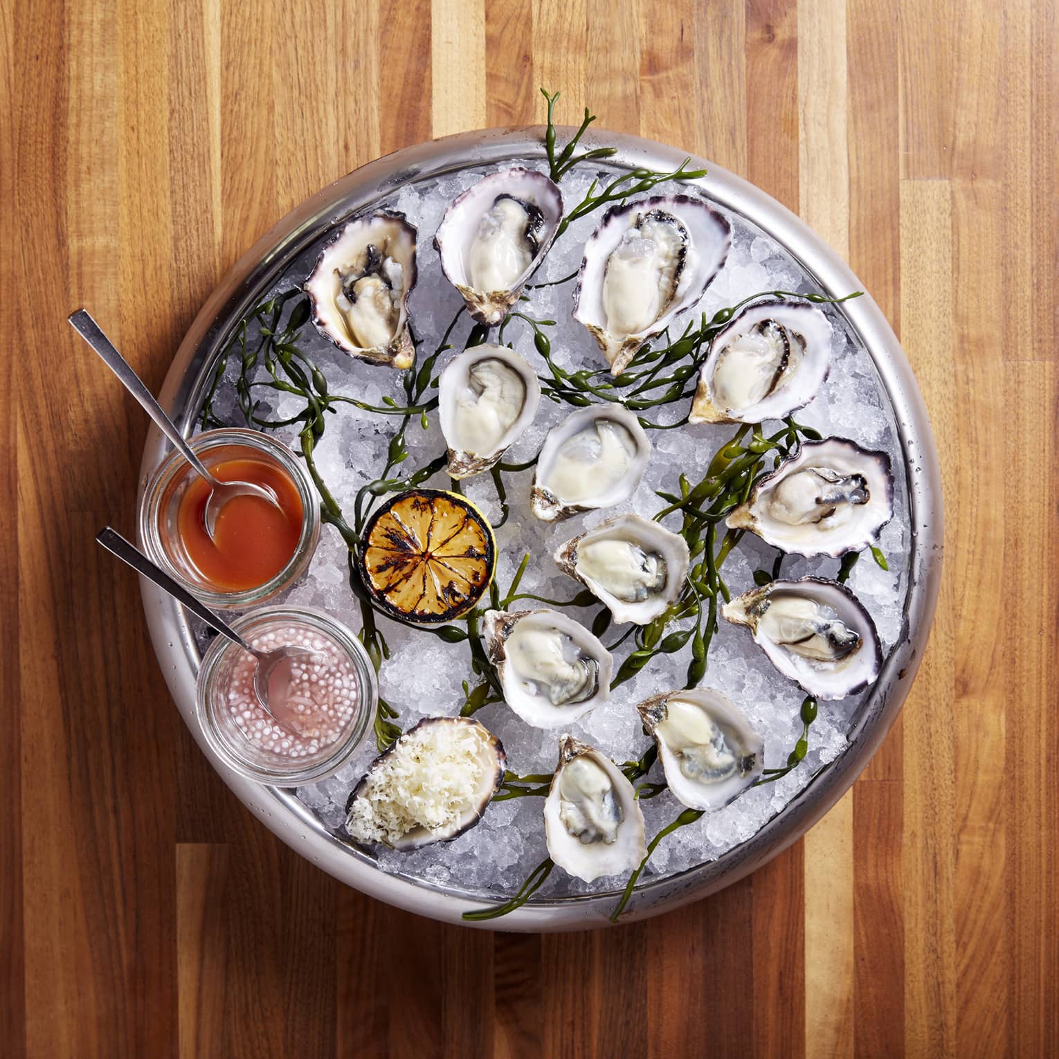 Aerial view of round silver platter with half shells and fresh oysters on ice, roasted lemon half and sauce bowls