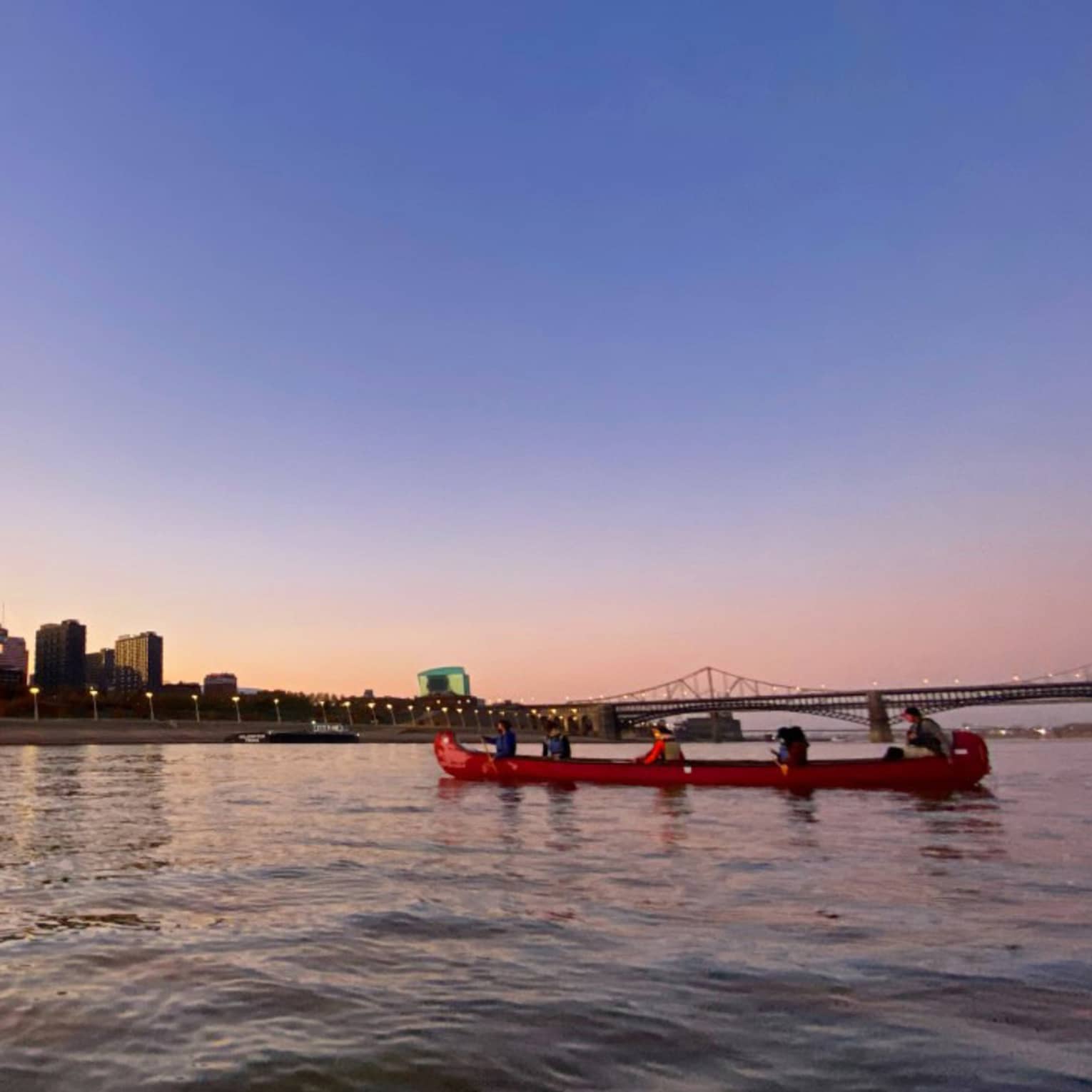 A group of people canoeing on a river at dusk, with a bridge, Gateway Arch, and city skyline visible in the background under a clear sky 
