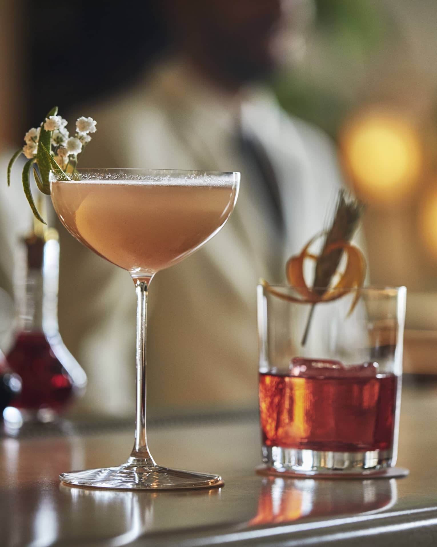 Two cocktails with garnish sit on a bar top with bottles and blurred image of man in background 