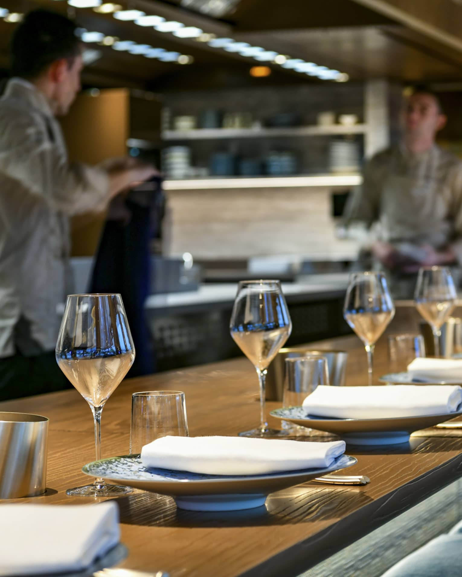 Two restaurant staff interact behind wood-topped bar with elegant table settings lining it