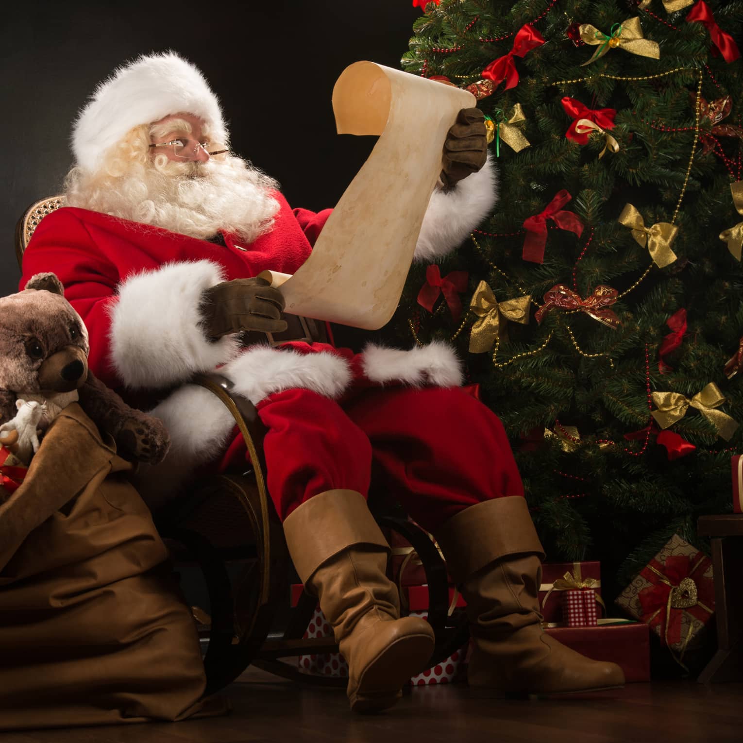 Santa Claus seated in large rocker holding a scroll, flanked by decorated Christmas tree with gifts and overflowing gift bag