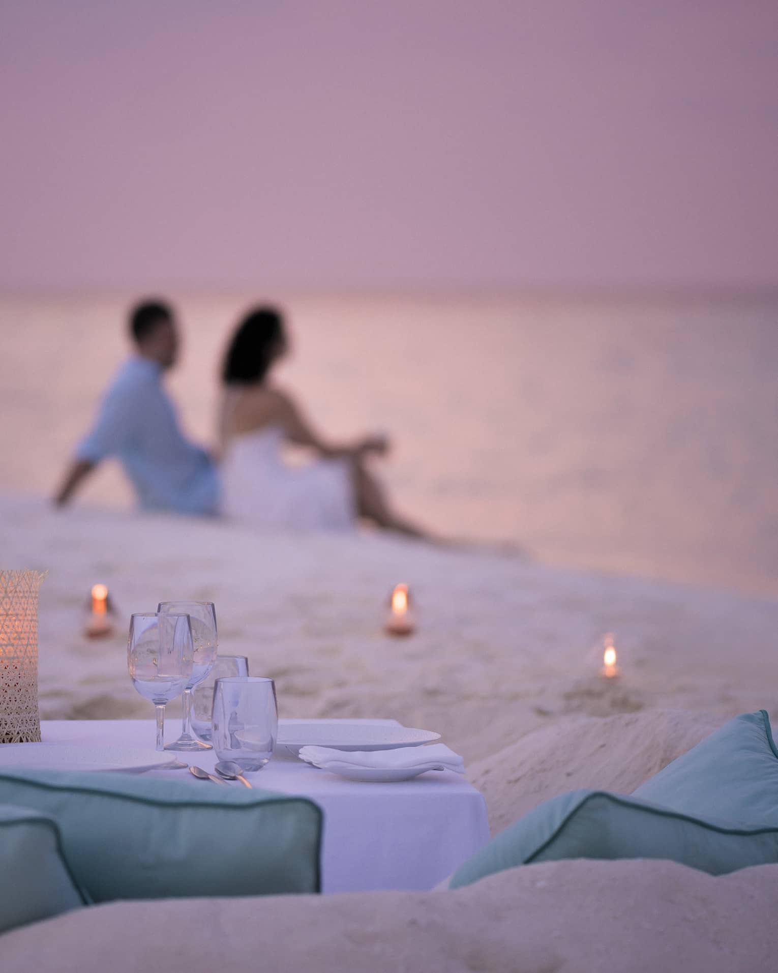 Private dining table in front of sand beach with tealight candles, couple sitting in sand