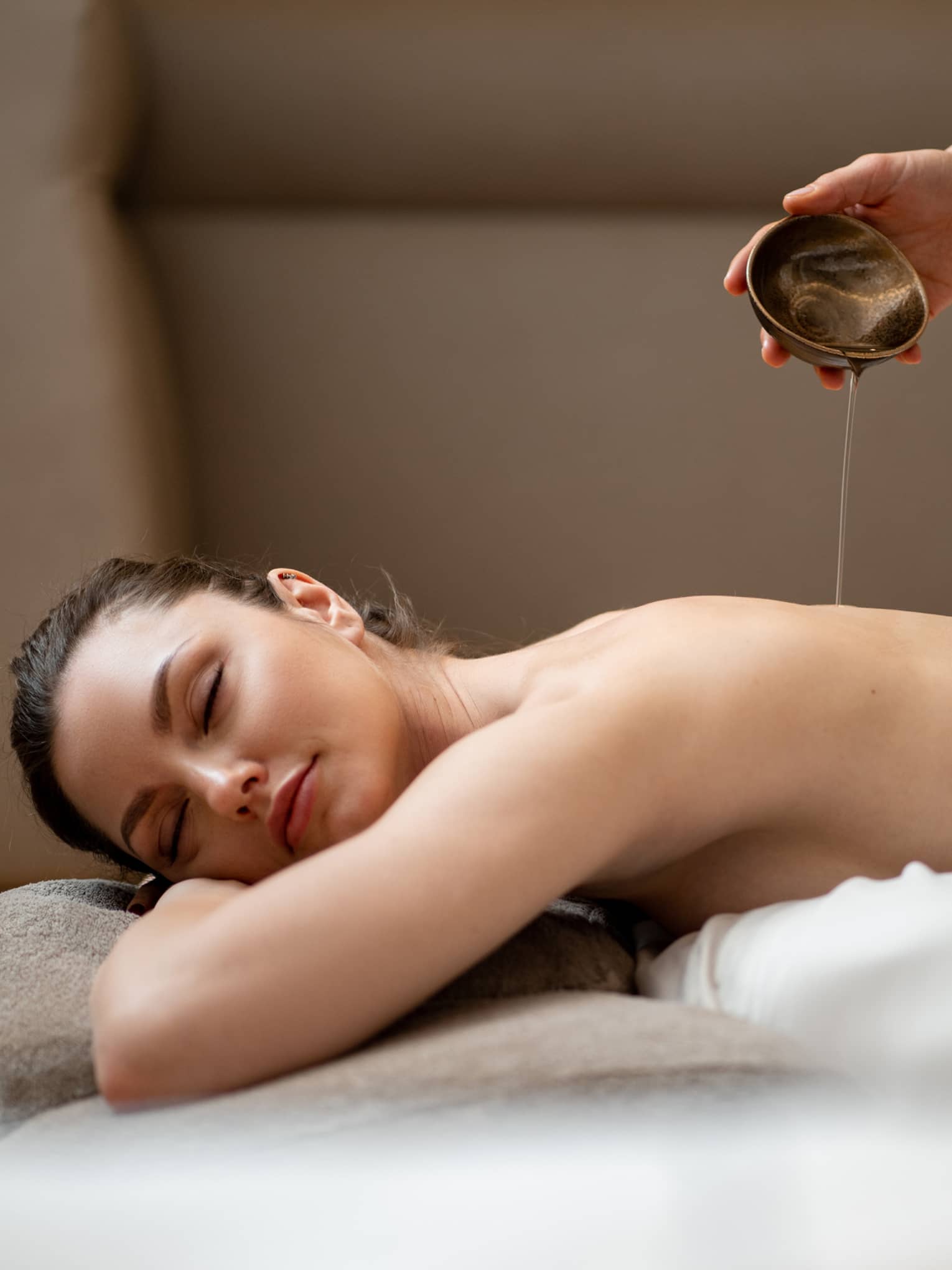 Woman lying with eyes closed on stomach while spa therapist pours oil from dish on her back