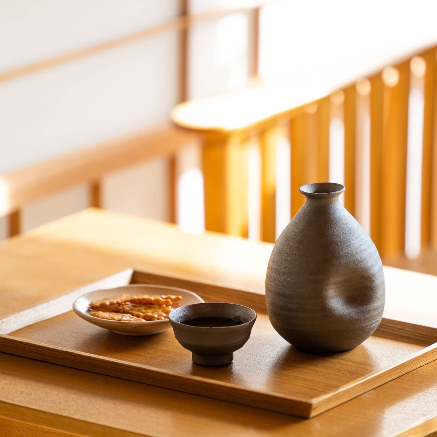 Close-up of a dark ceramic sake carafe with matching cup and a small plate of crackers on a wooden tray and table.