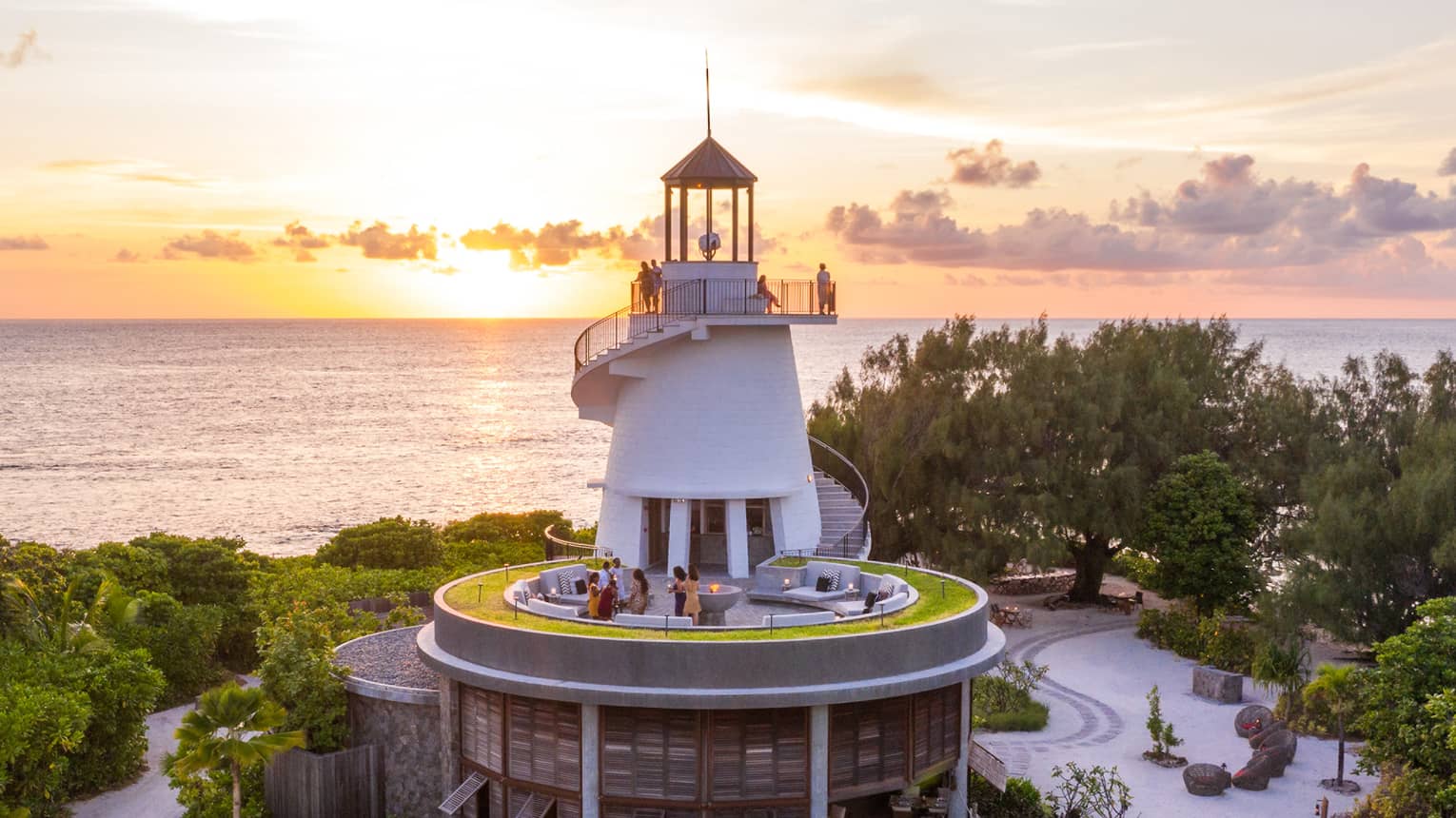 Lighthouse nestled among greenery with people on different levels, overlooking the ocean at sunset