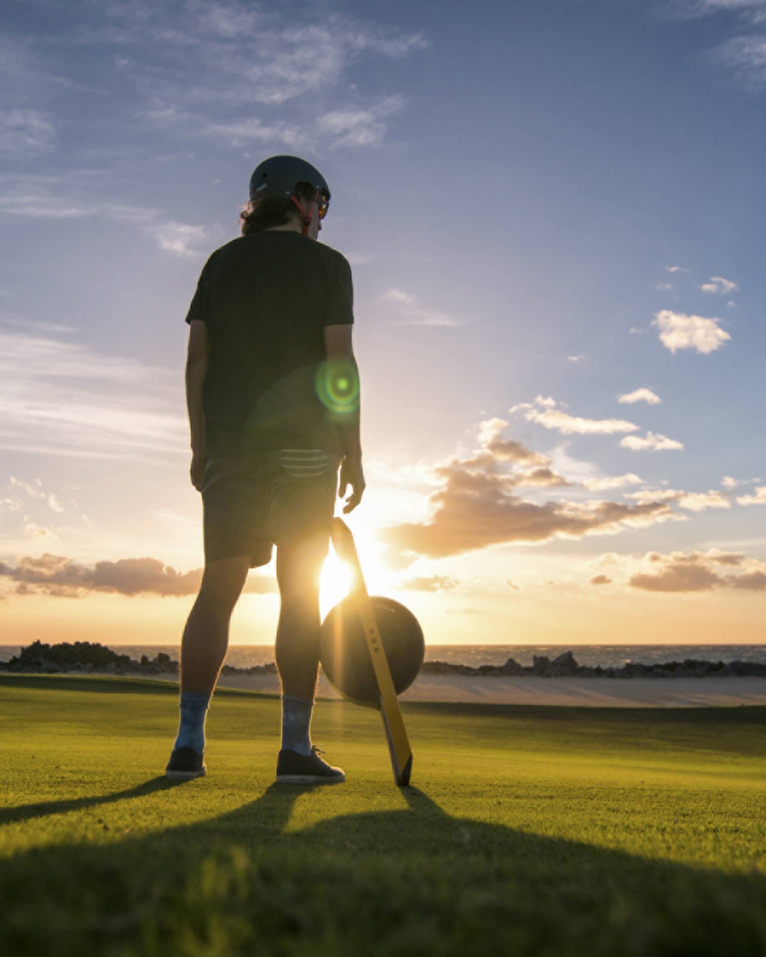 A young boy standing on a large field of green grass with the sun setting in the distance in front of him.
