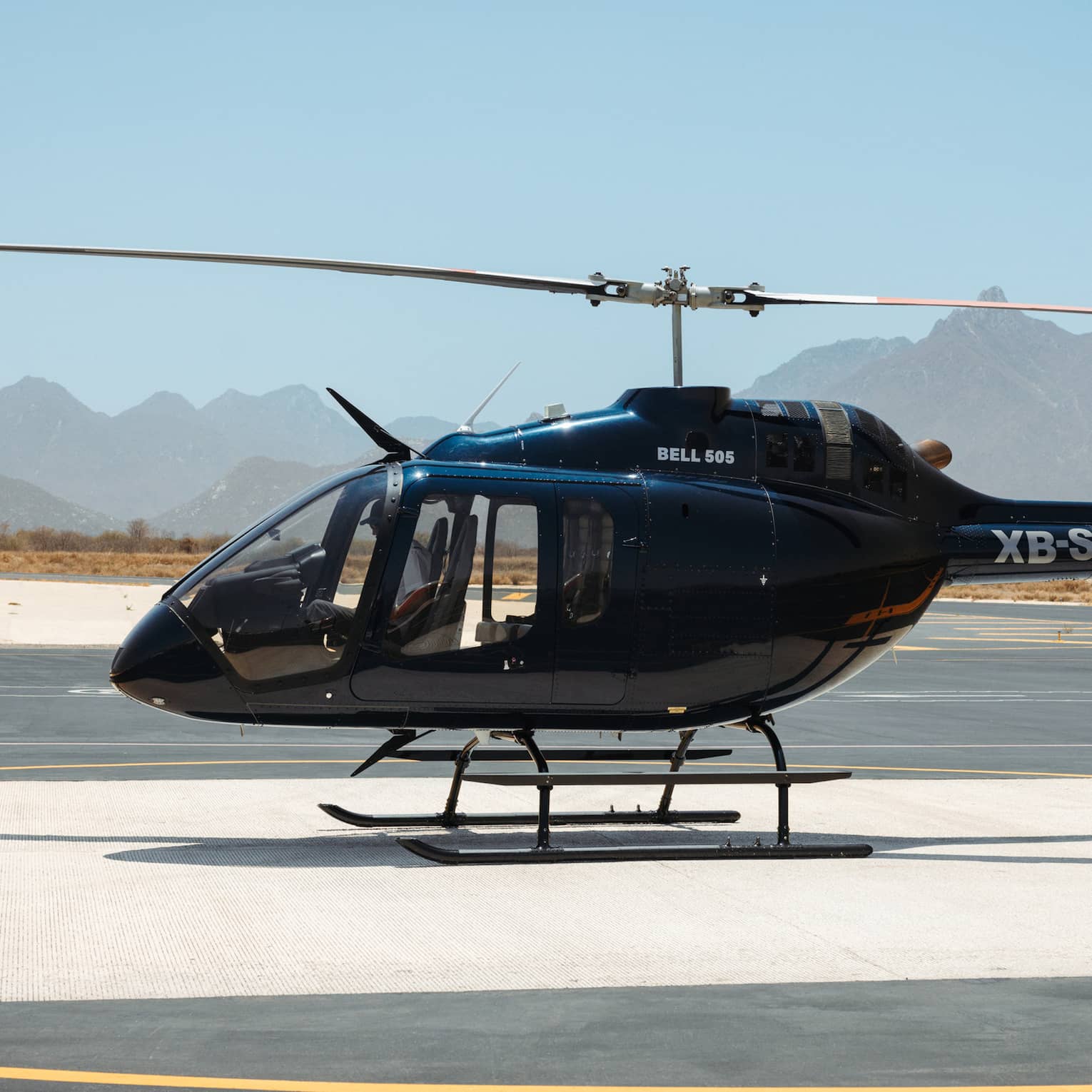 Side view of a piloted black helicopter parked on a runway, tall golden grass and hazy jagged mountains in the distance.