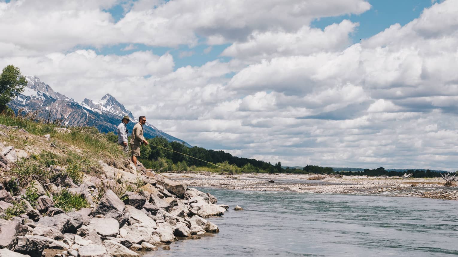 Two men fish on the rocky bank of a lake with fluffy white clouds above them in the sky