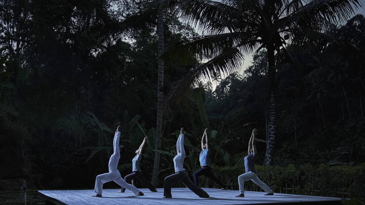 Five people kneel in yoga pose with arms above head on wood platform in forest at night