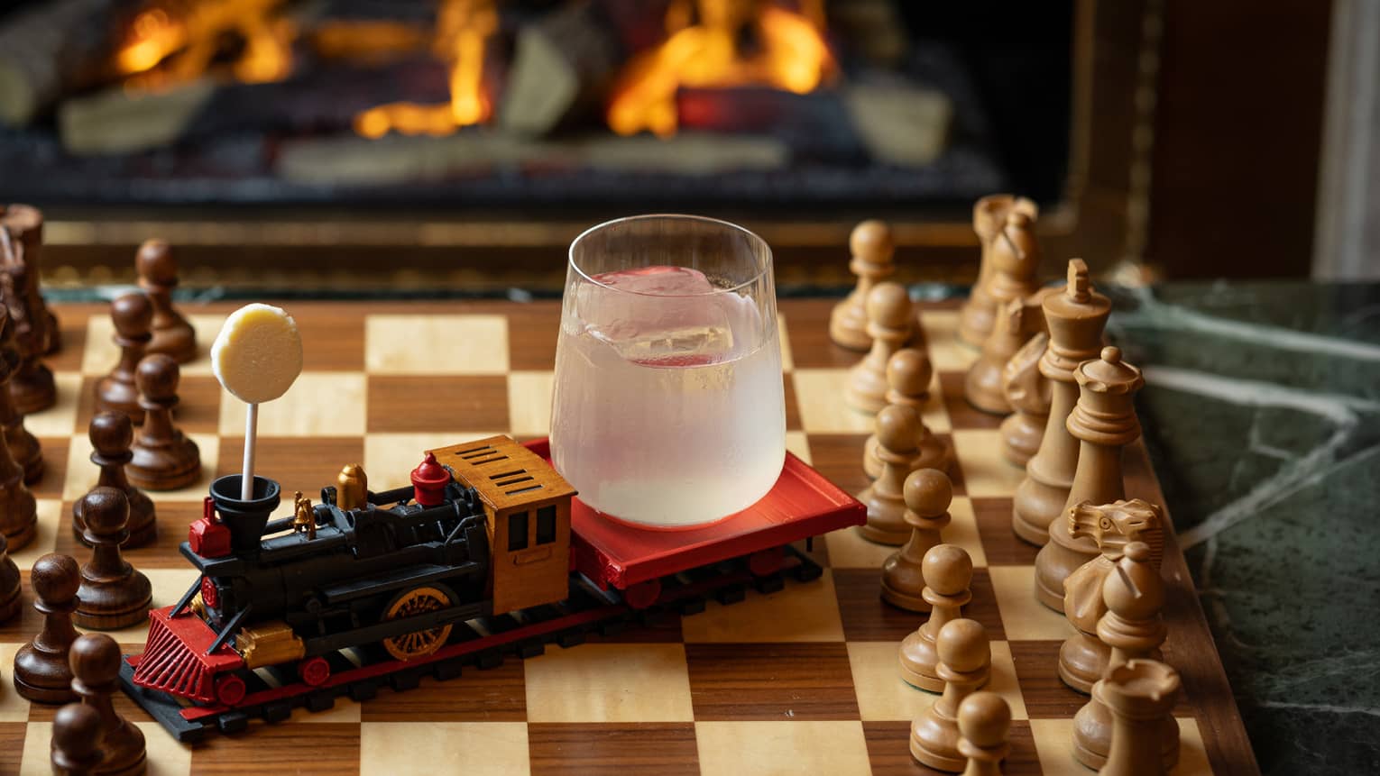 A clear tumbler holding an opaque whitish liquid sits on the back of a model train engine, which is on a chessboard on a tabletop, with a fireplace in the background.