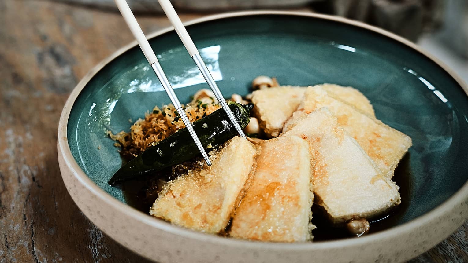 A person uses chopsticks to select a piece of tofu from a colourful glazed bowl.