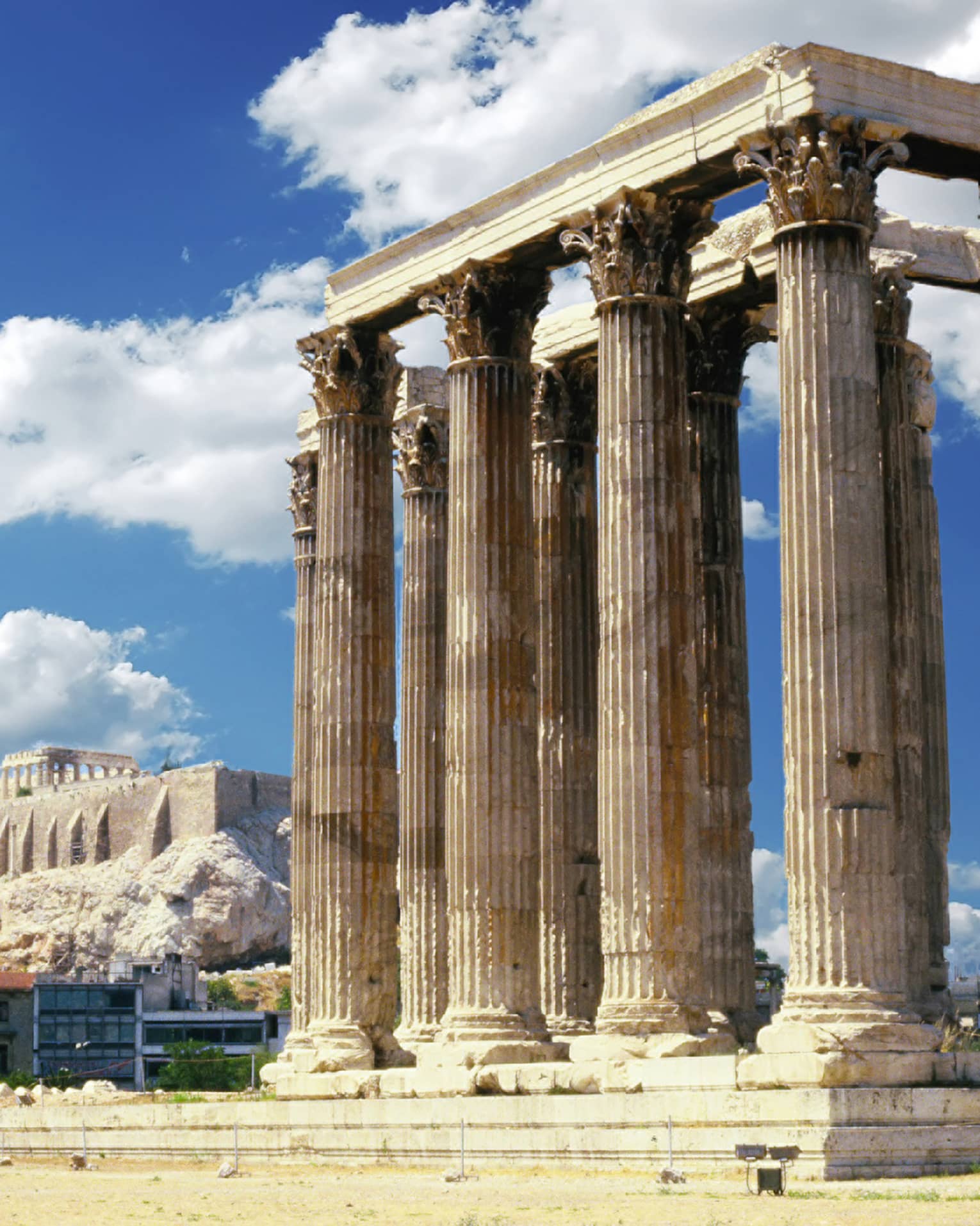 A temple with towering columns featuring intricate designs; more ruins and the Parthenon prominent in the background.
