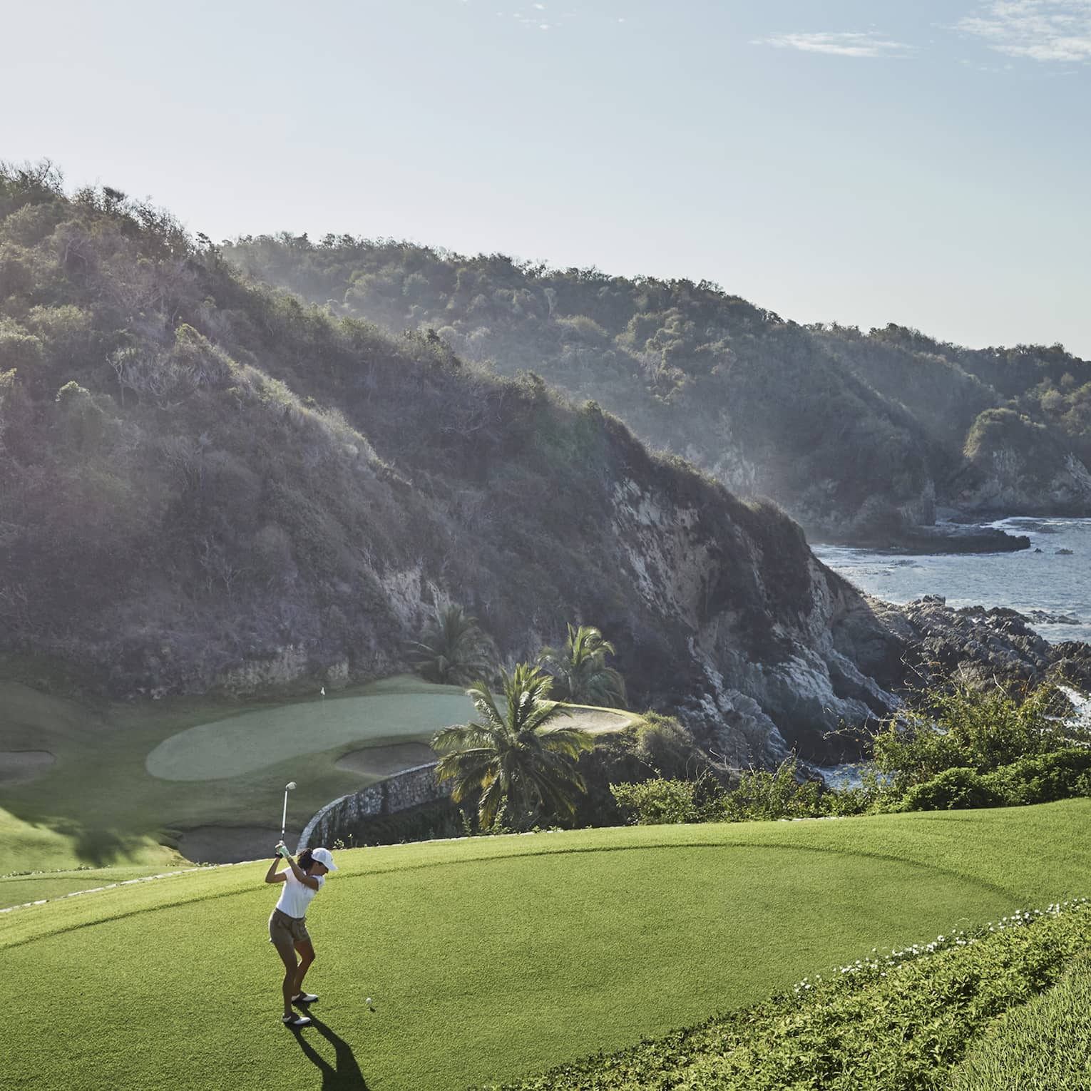 Golfer in mid-backswing, ready to strike toward a distant green at the base of an immense forested hill bordering the ocean. 