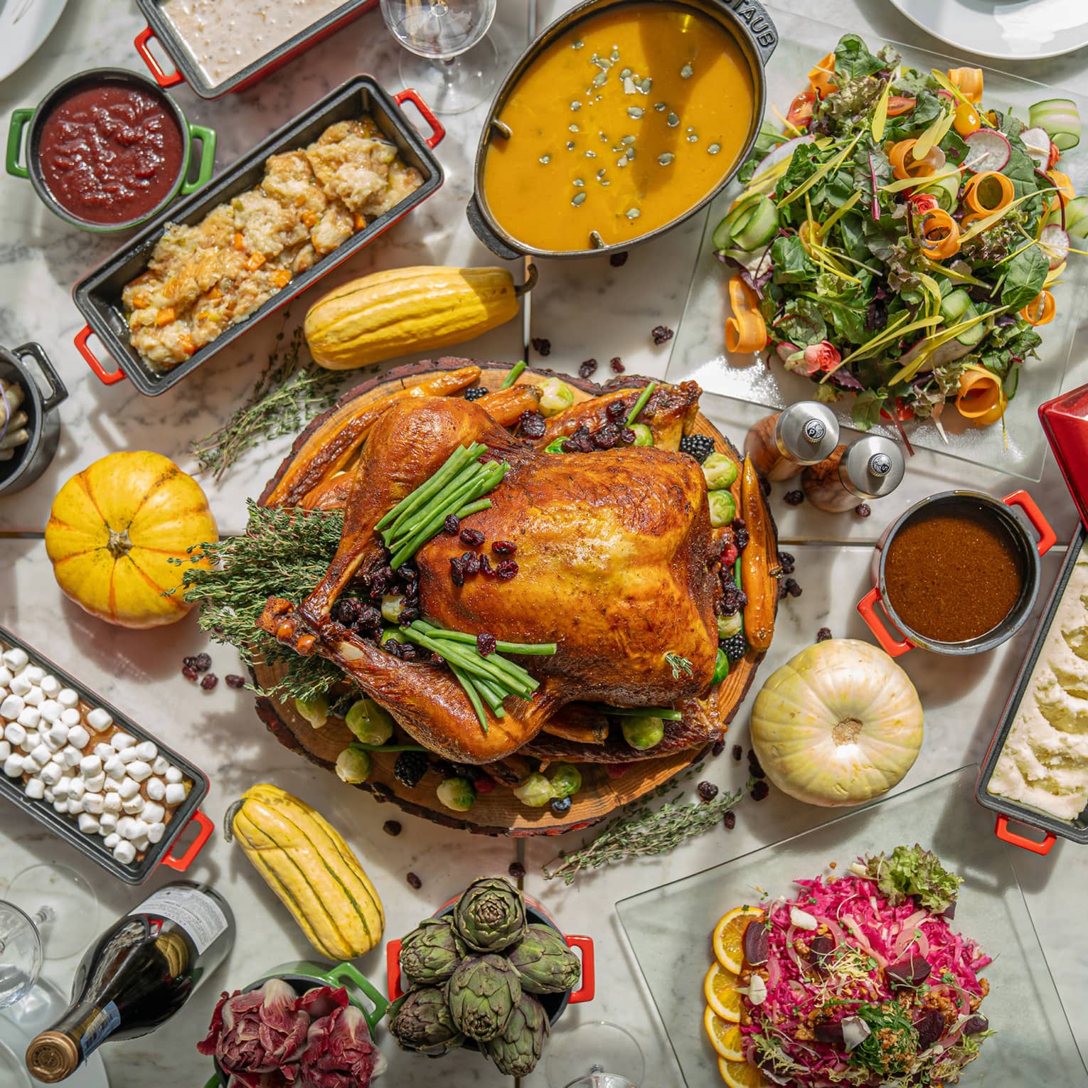 Whole roasted turkey sits in the middle of a white marble table surrounded by a variety of colorful side dishes, salads and desserts