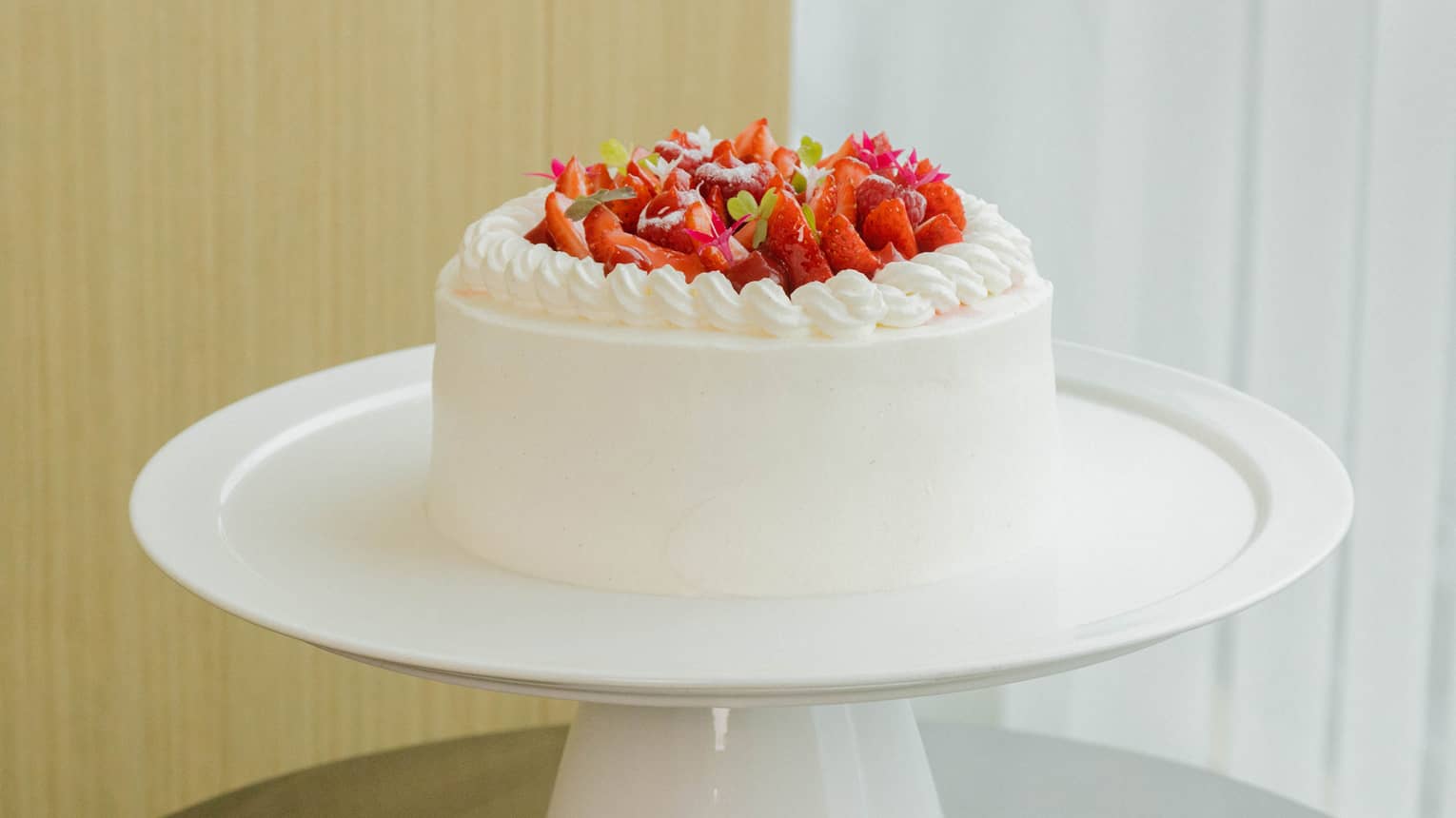 Round strawberry shortcake with white icing and topped with fresh sliced strawberries is set atop a round white cake stand