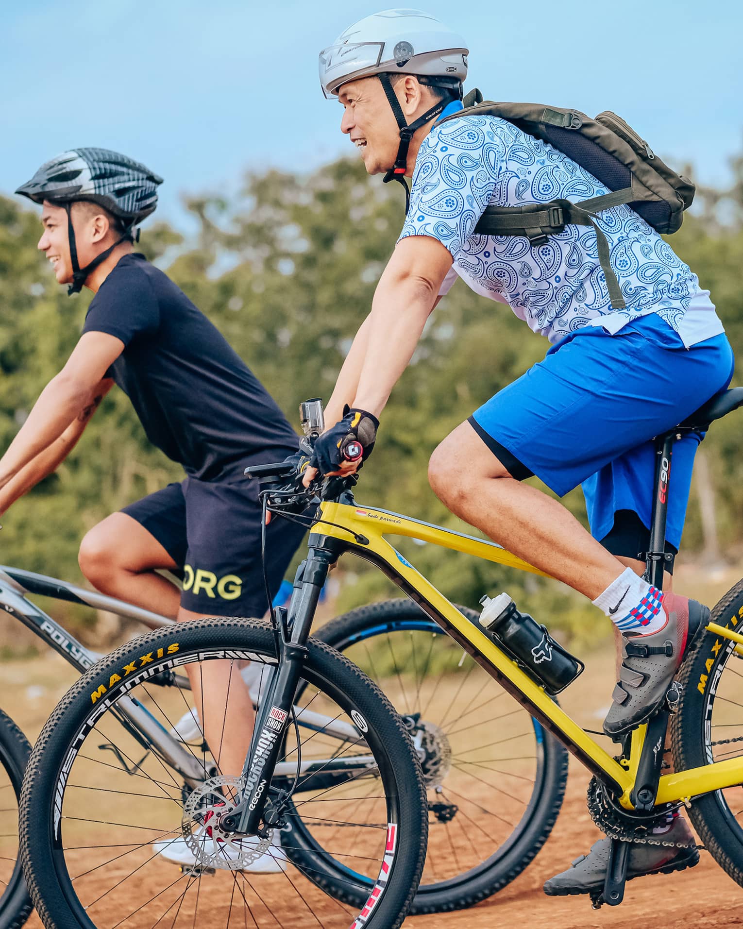 Side view of two smiling mountain bikers in helmets as they coast down a gentle slope on a dirt path alongside trees.