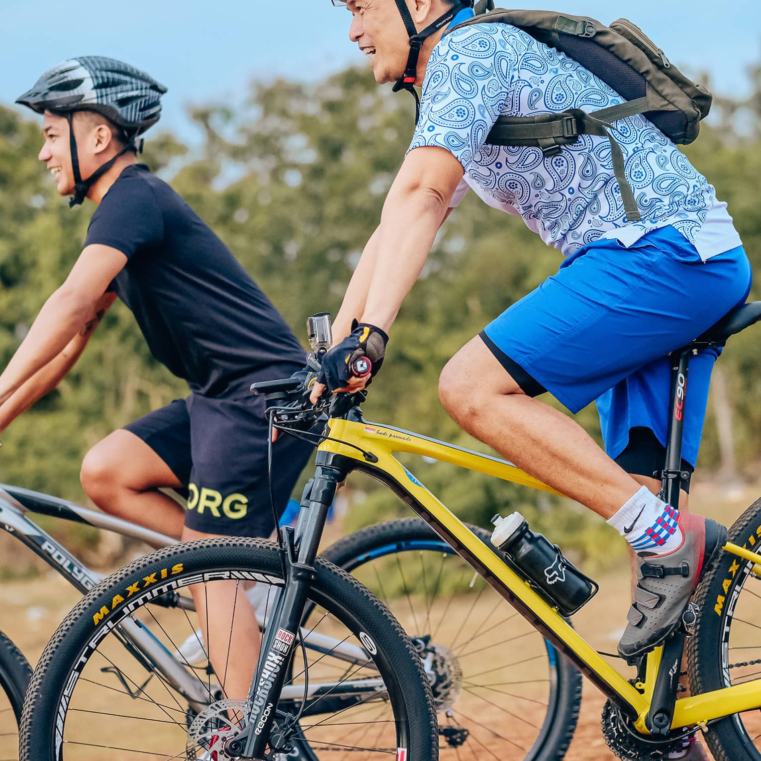 Side view of two smiling mountain bikers in helmets as they coast down a gentle slope on a dirt path alongside trees.