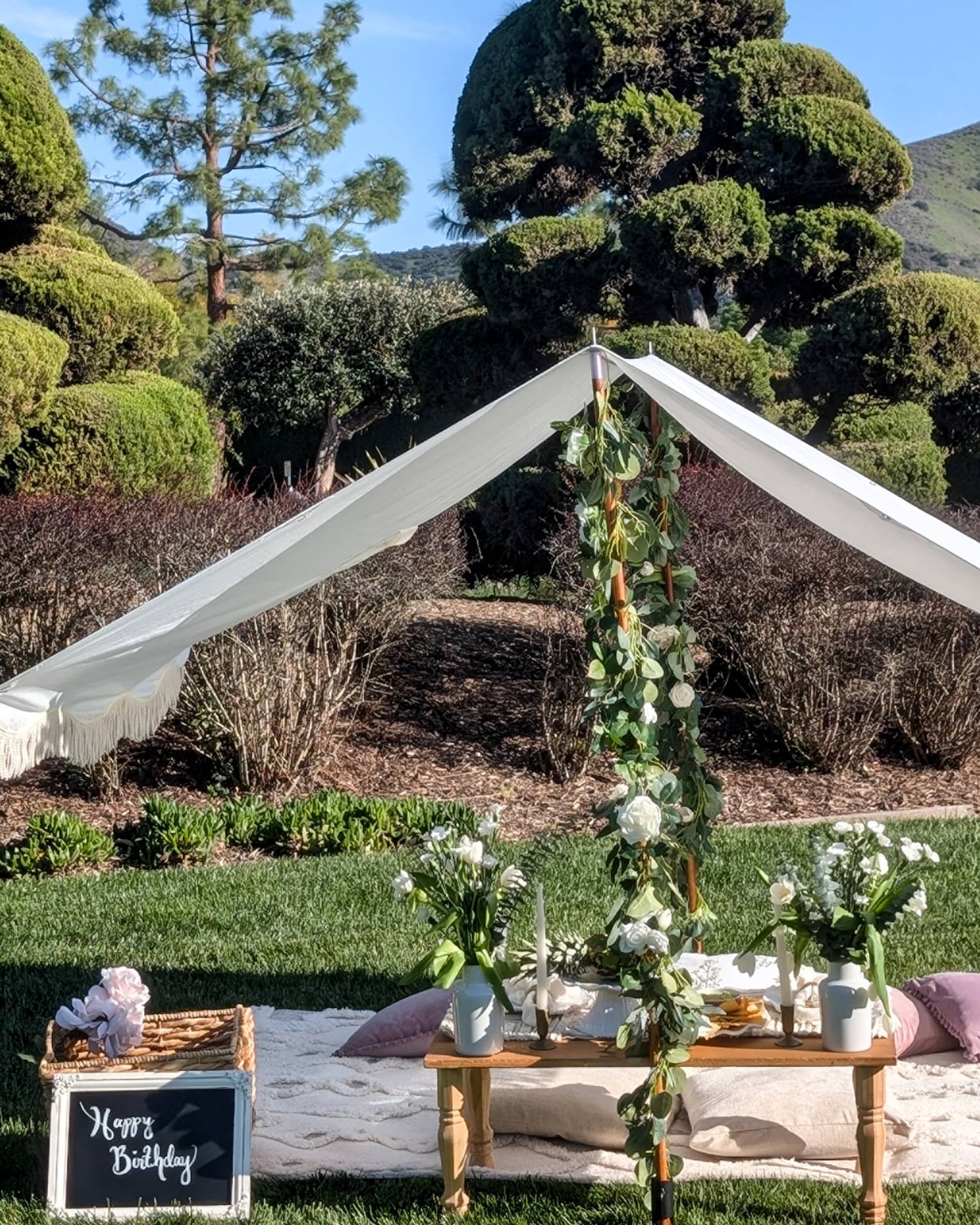 Picnic set up on a lawn with blanket, pillows, small table, flower garlands and white canopy