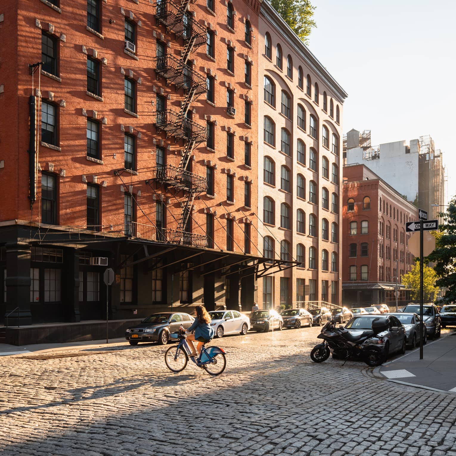 A person riding a bicycle in front of of a brick building with cars lining the street.