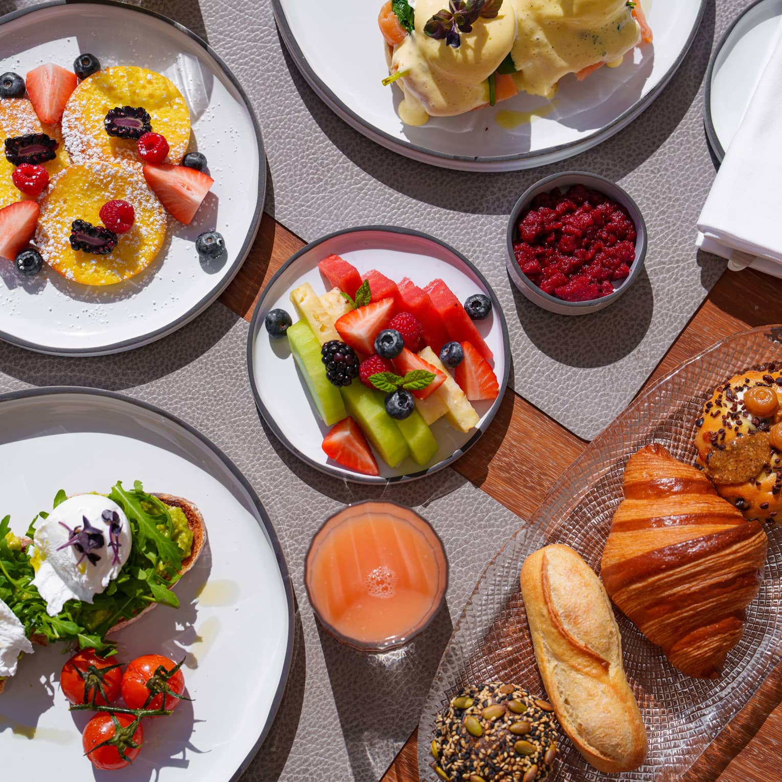 A plate of eggs Benedict next to three tomatoes on a vine, a plate of fresh fruit, a plate of pancakes and fresh berries, a platter of pastries, a bowl of yogurt and granola and a glass of juice fill a dining table