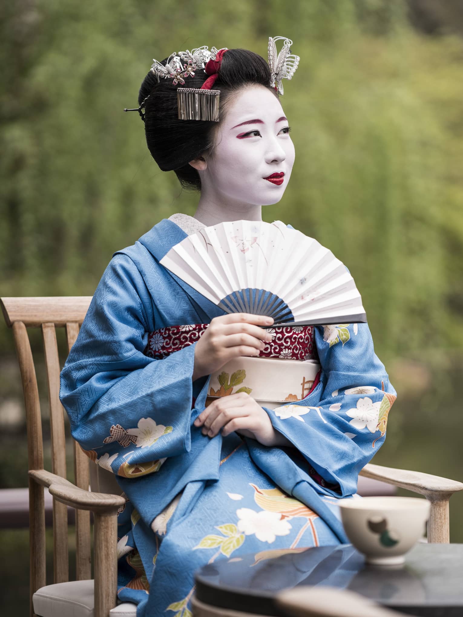 Geiko, a traditional Japanese entertainer, dressed in kimono and holding fan, sitting with cup of tea in garden