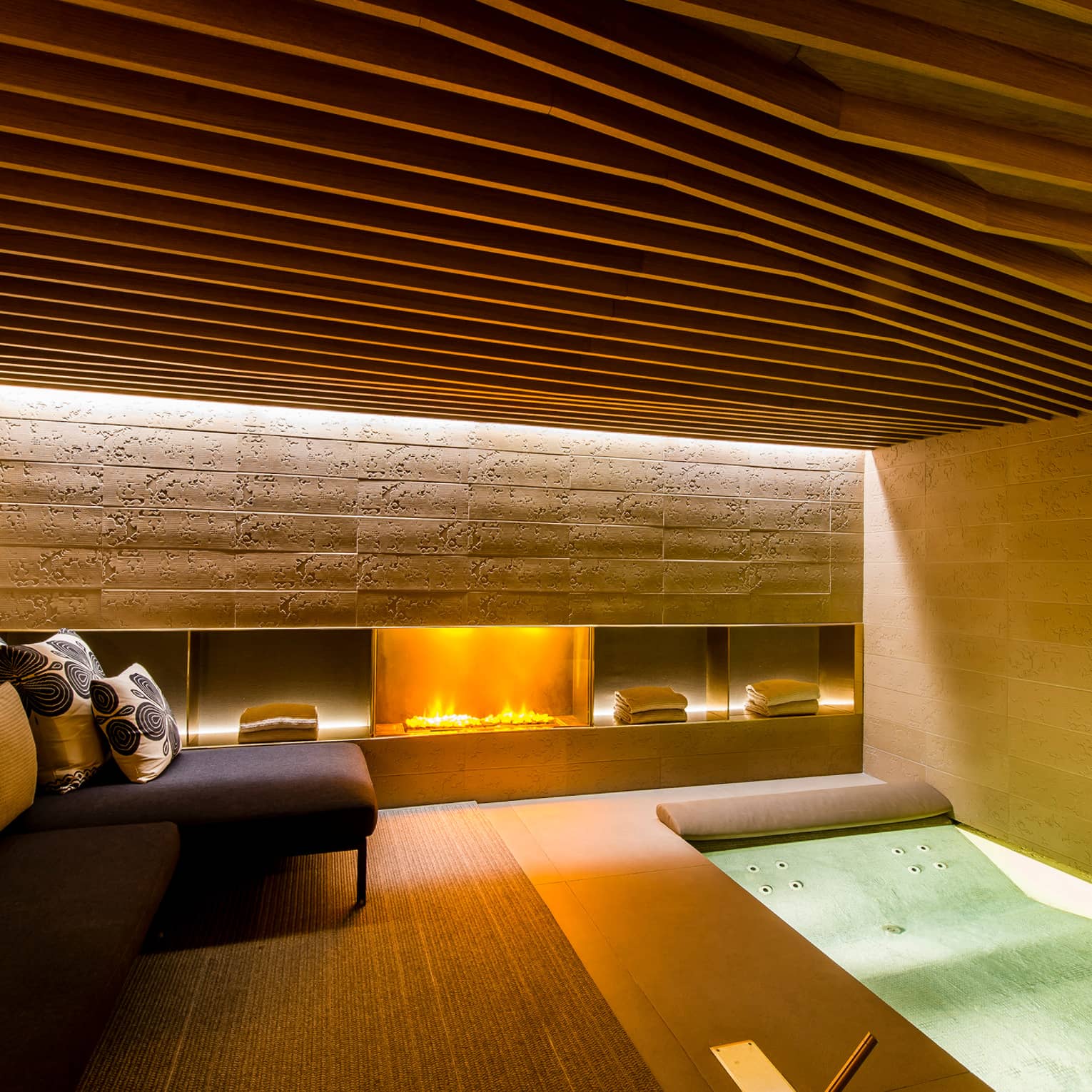 Gas fireplace in under stone wall in Urban Spa, small L-shaped sofa, cushions, spa tub
