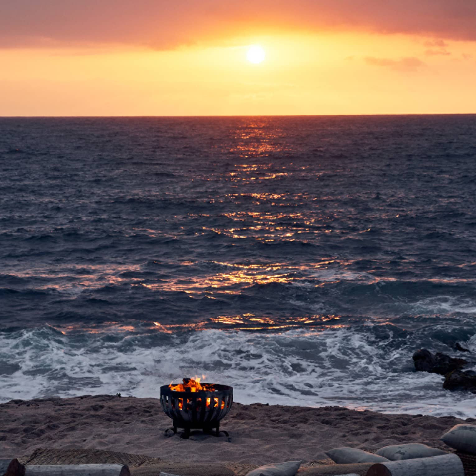 Fire burns in an iron pit on the beach, semi-encircled by driftwood log seats facing the sun setting into the ocean horizon.