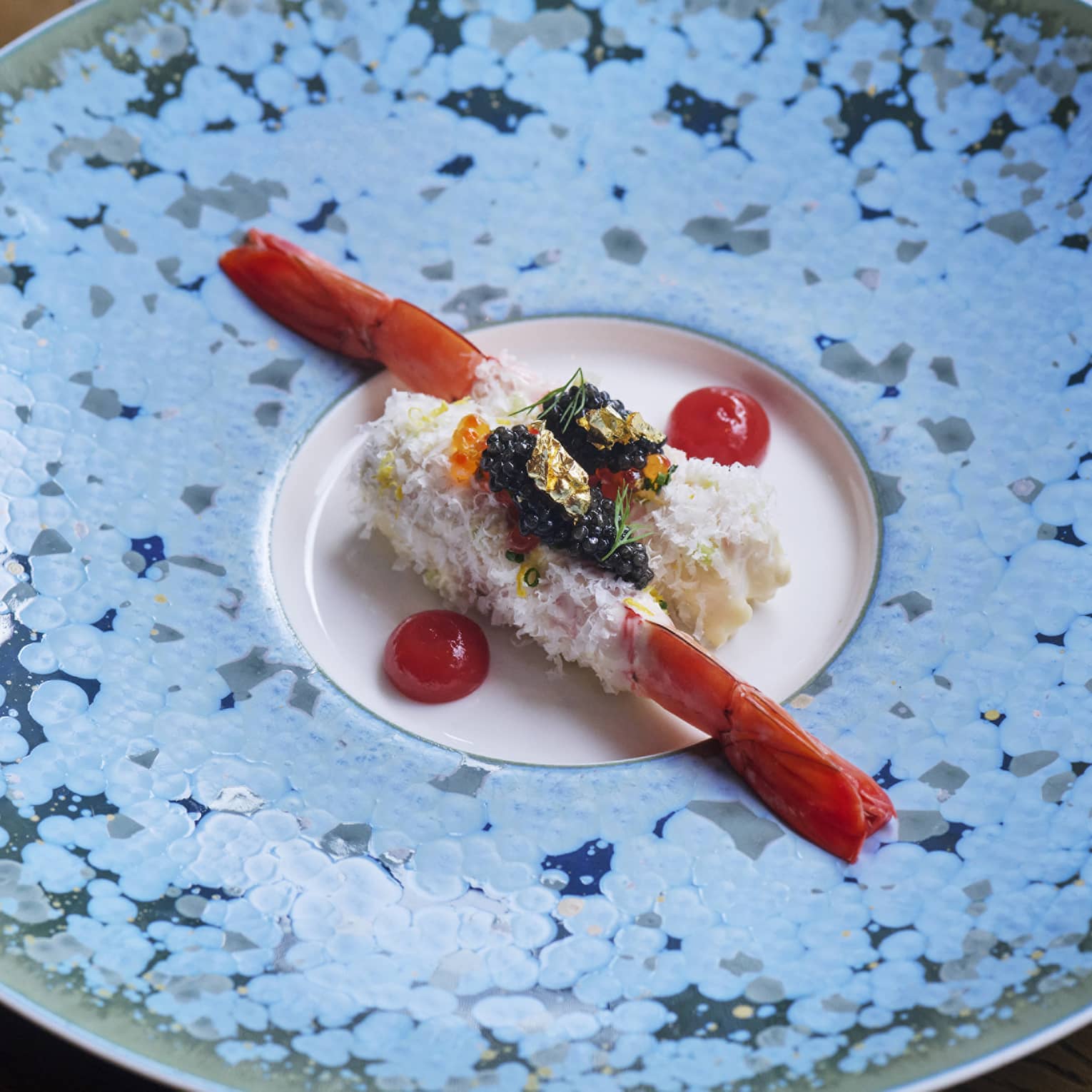 Two elegantly plated prawns topped with garnishes and flanked by cherry tomato halves in centre of blue speckled plate