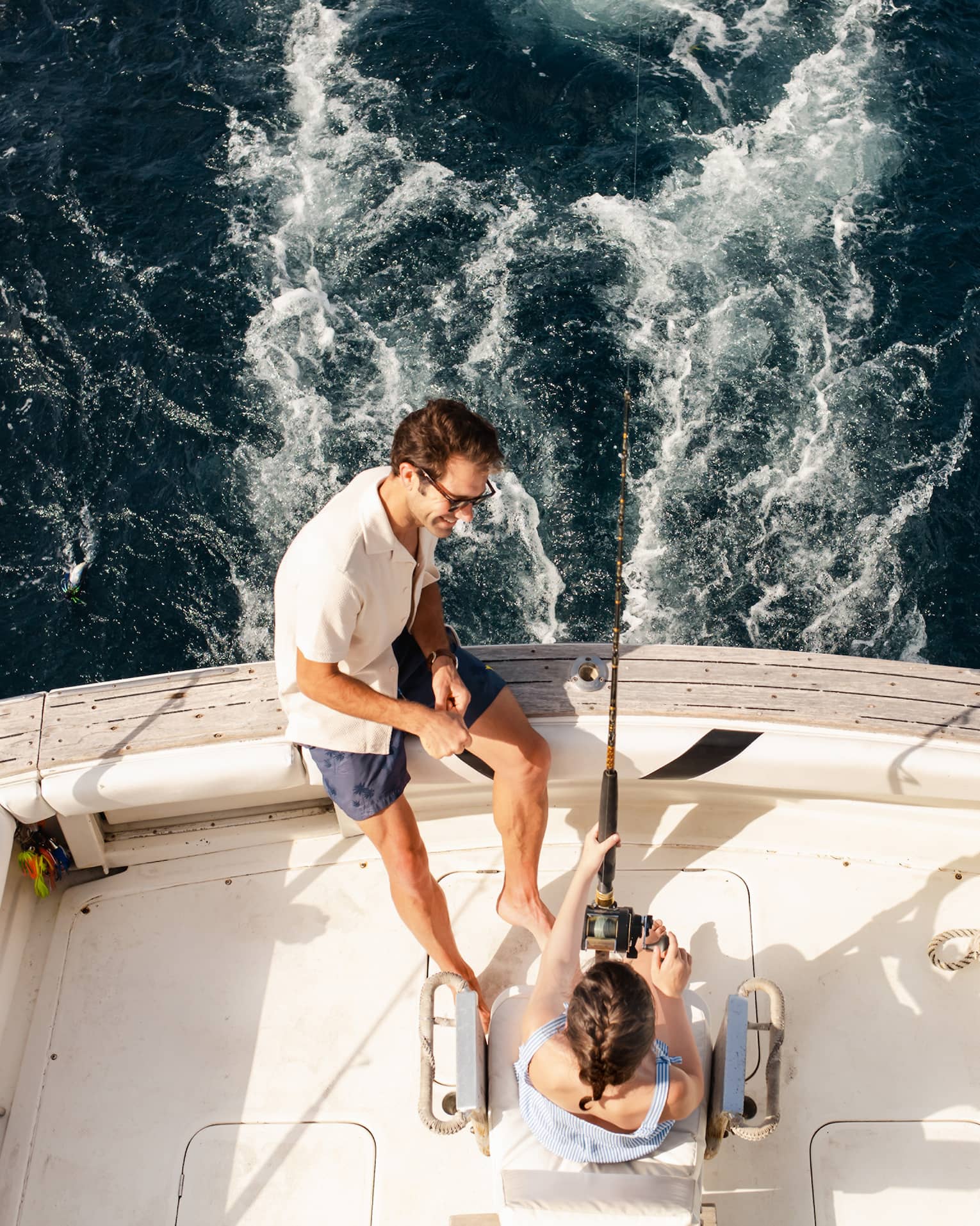 Two people sit at the back of a fishing boat, with one person holding a fishing rod and the other sitting close by on the edge of the boat