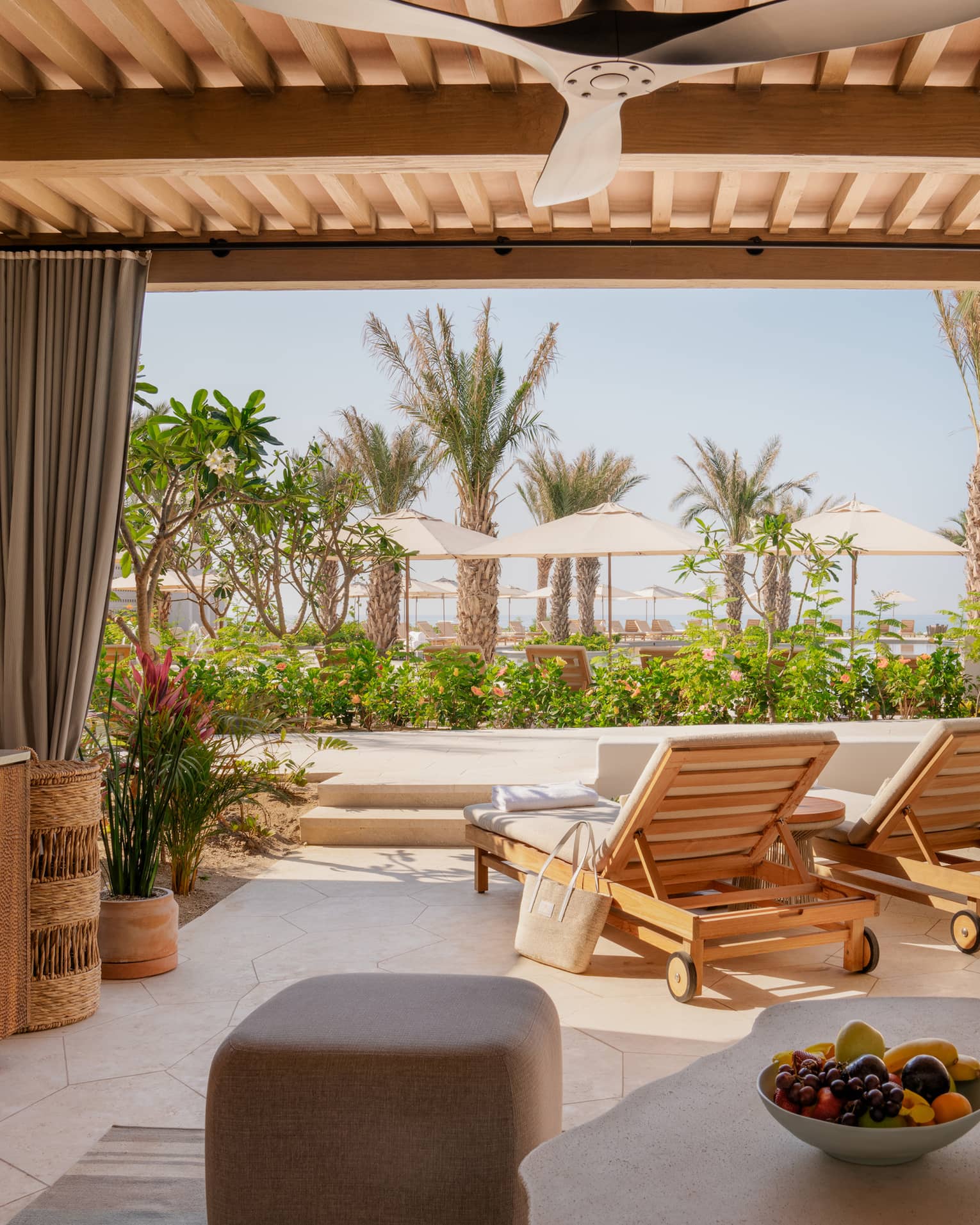 View from inside a neutral cabana with champagne, a fruit bowl and lounge chairs facing out towards palm trees and umbrellas.