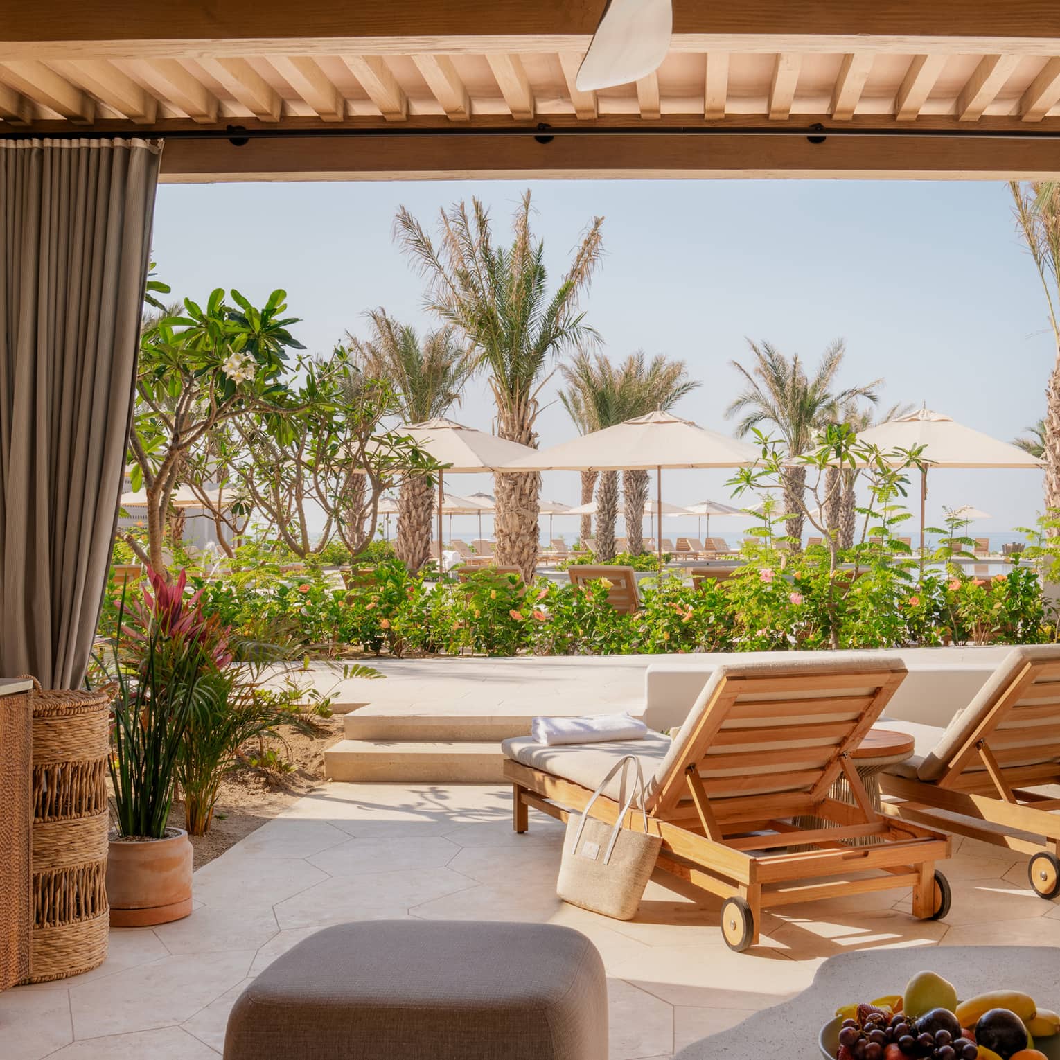 View from inside a neutral cabana with champagne, a fruit bowl and lounge chairs facing out towards palm trees and umbrellas.