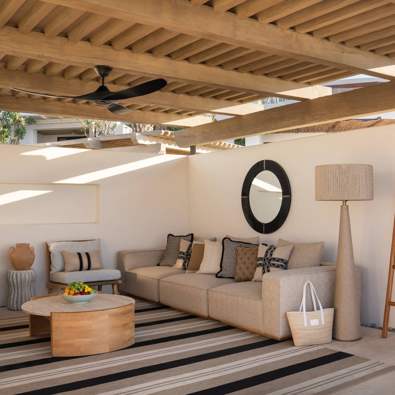 A serene cabana with a raised wooden roof and plaster walls. It's filled with comfortable furniture and a large striped rug.