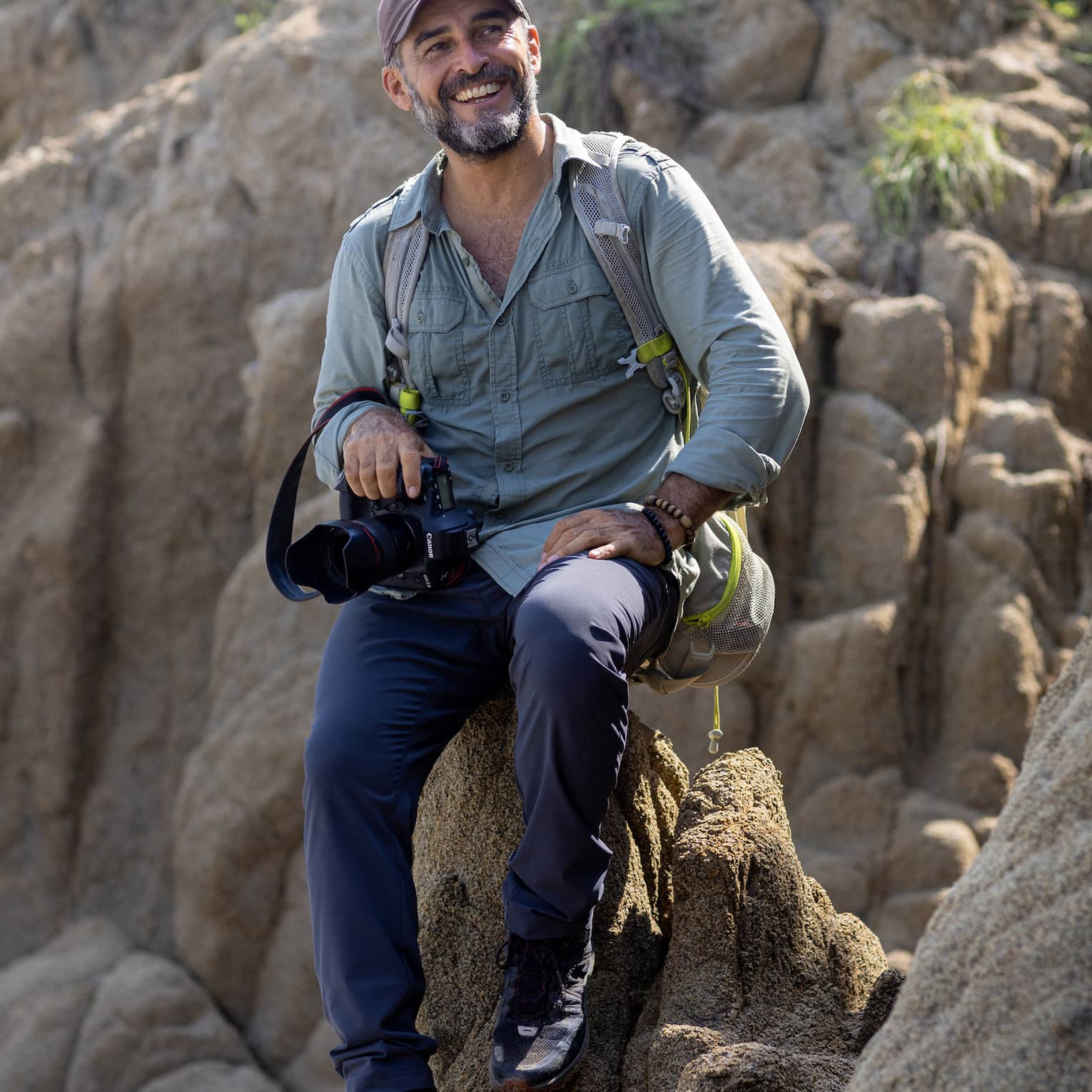 A photographer wearing a backpack and holding a camera in one hand sits atop a rock formation, smiling into the distance.
