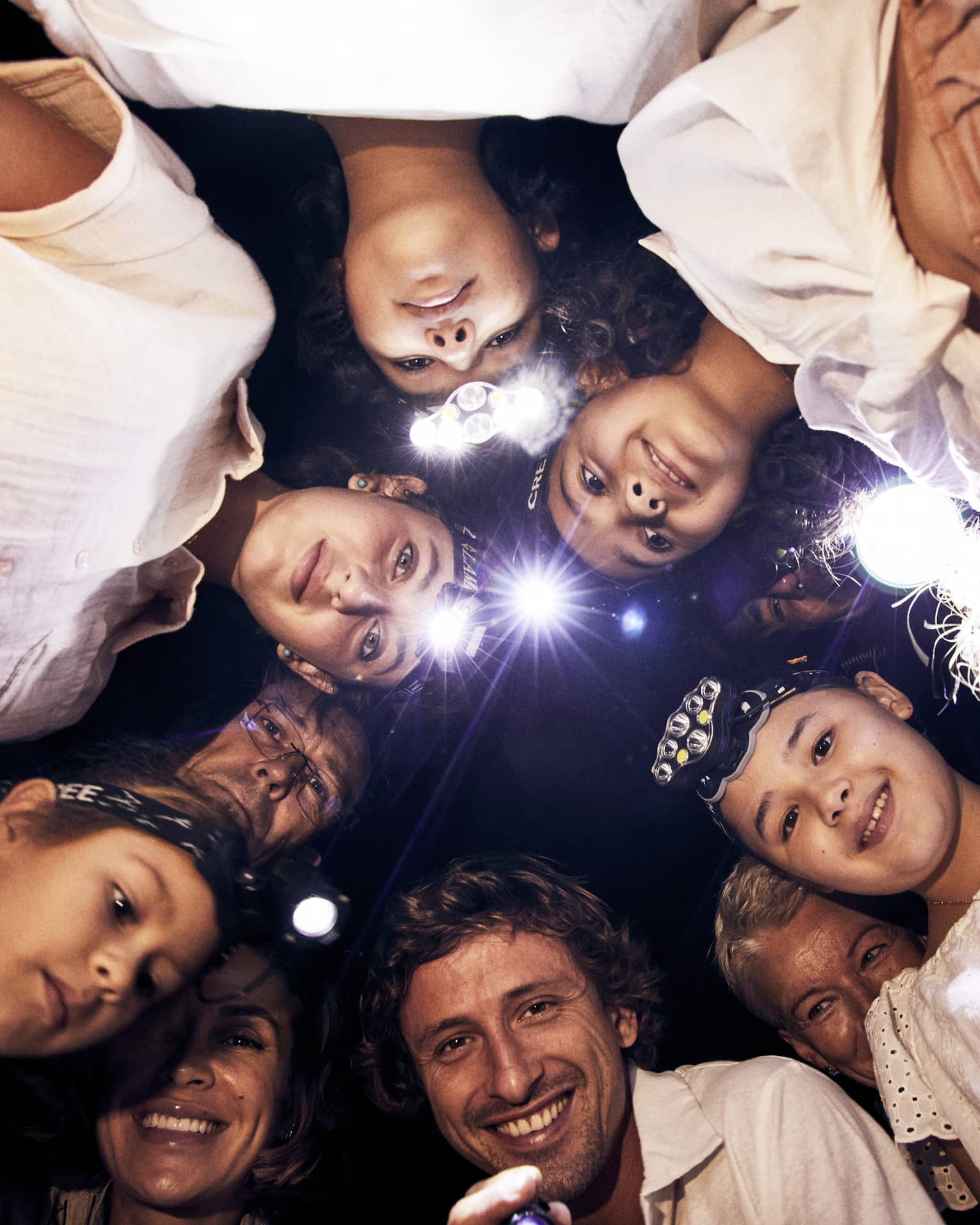 A group of children and adults, some wearing headlamps, stand in a circle and smile downwards into the camera. 