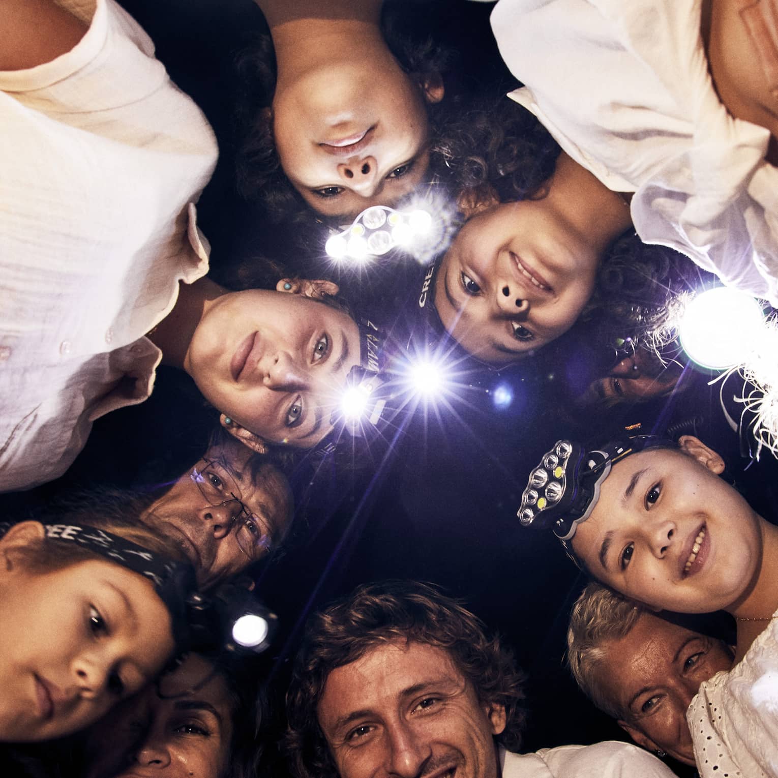 A group of children and adults, some wearing headlamps, stand in a circle and smile downwards into the camera. 