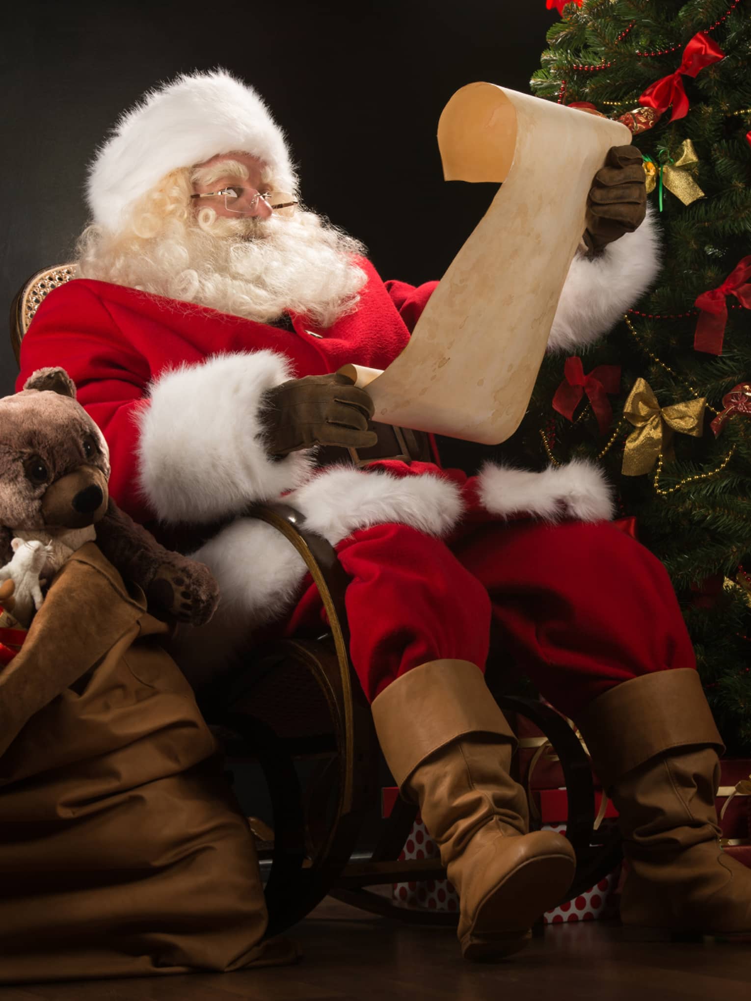 Santa Claus seated in large rocker holding a scroll, flanked by decorated Christmas tree with gifts and overflowing gift bag