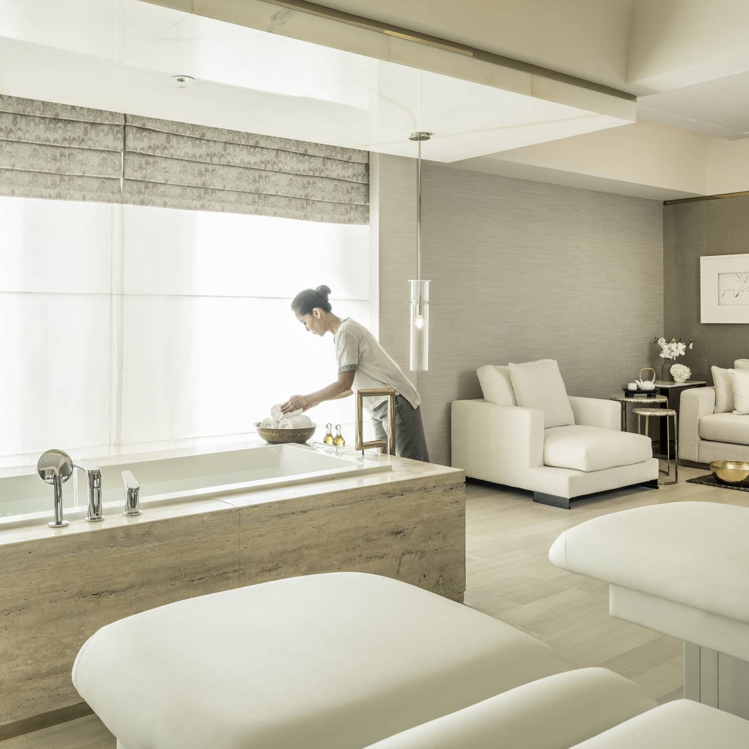 A spa attendant arranges some products in the spa suite, with two massage beds and a soaking tub in front