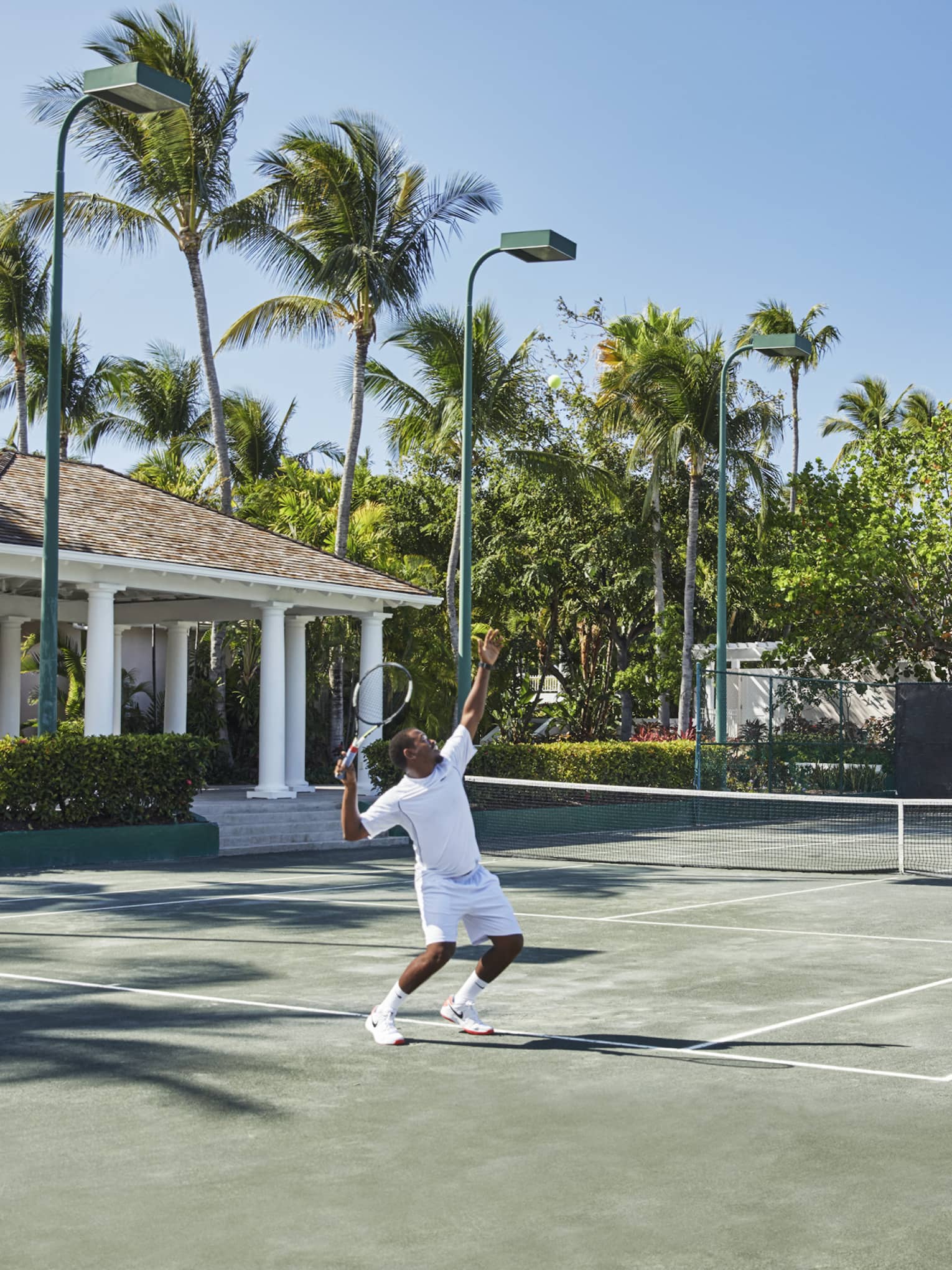 A man in all white trows a tennis ball high into the air, mid-serve 