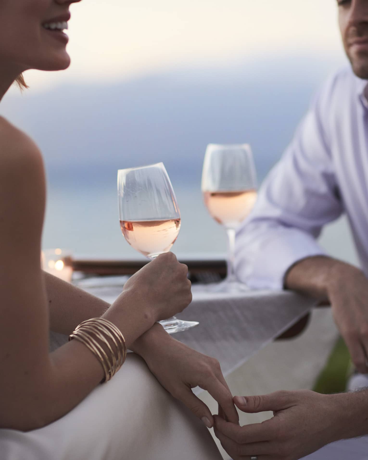 Close-up of woman and man in white, sitting and holding glasses of rose wine at dusk