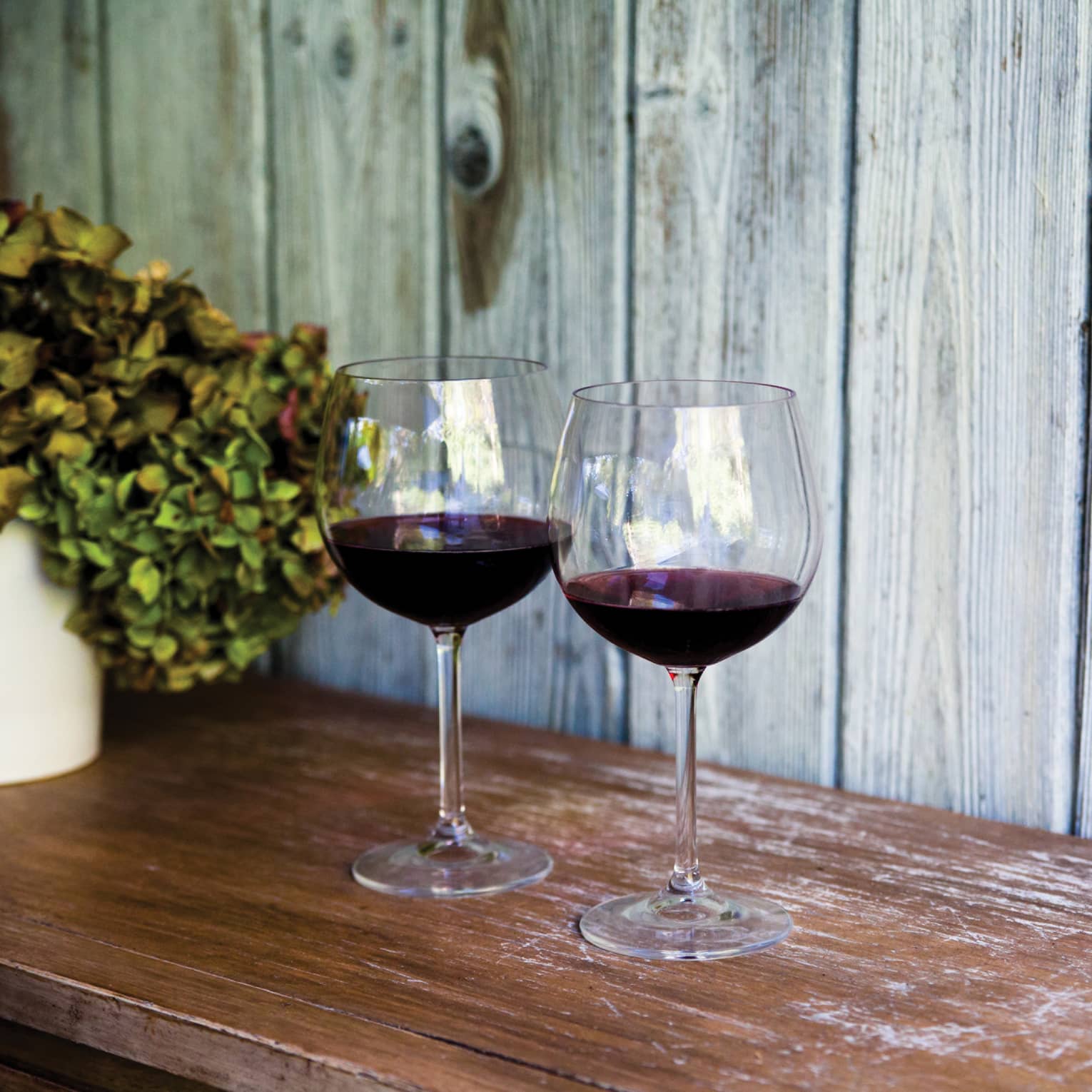 Two glasses of red wine and sit on wooden table, with white vase filled with hydrangeas 