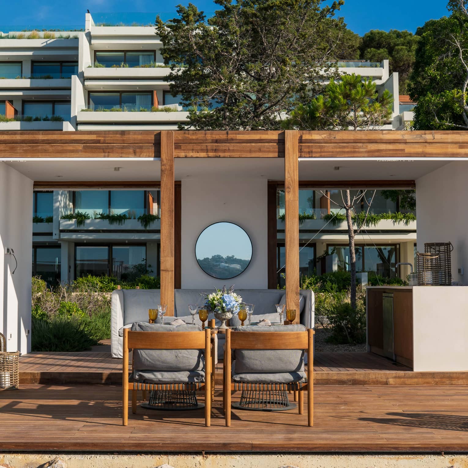 A pristine white cabana with wood accents frames a patio dining area in front of hotel balconies laden with greenery. 