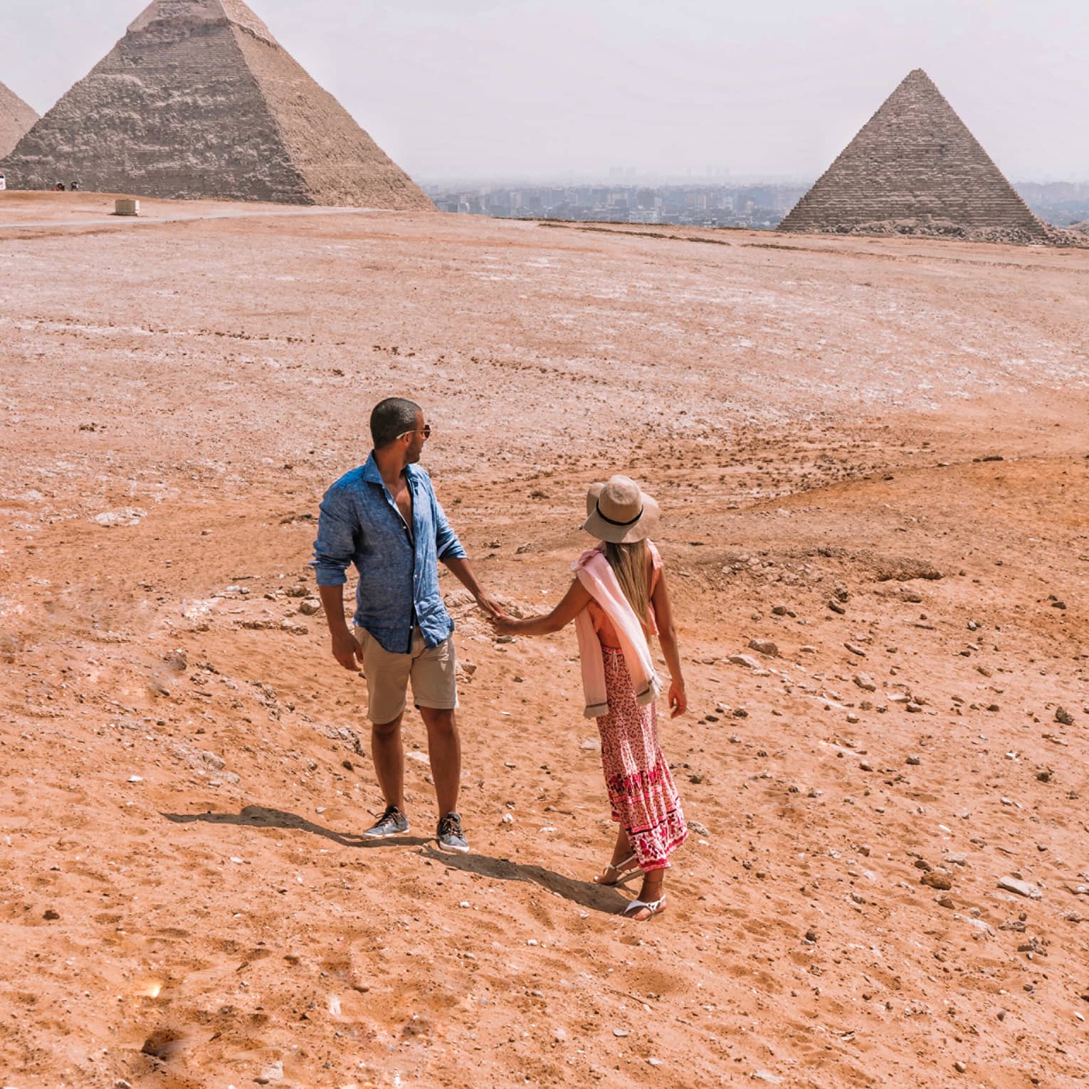 A couple holding hands turn back to look across the arid desert at the giant pyramids in the distance. 