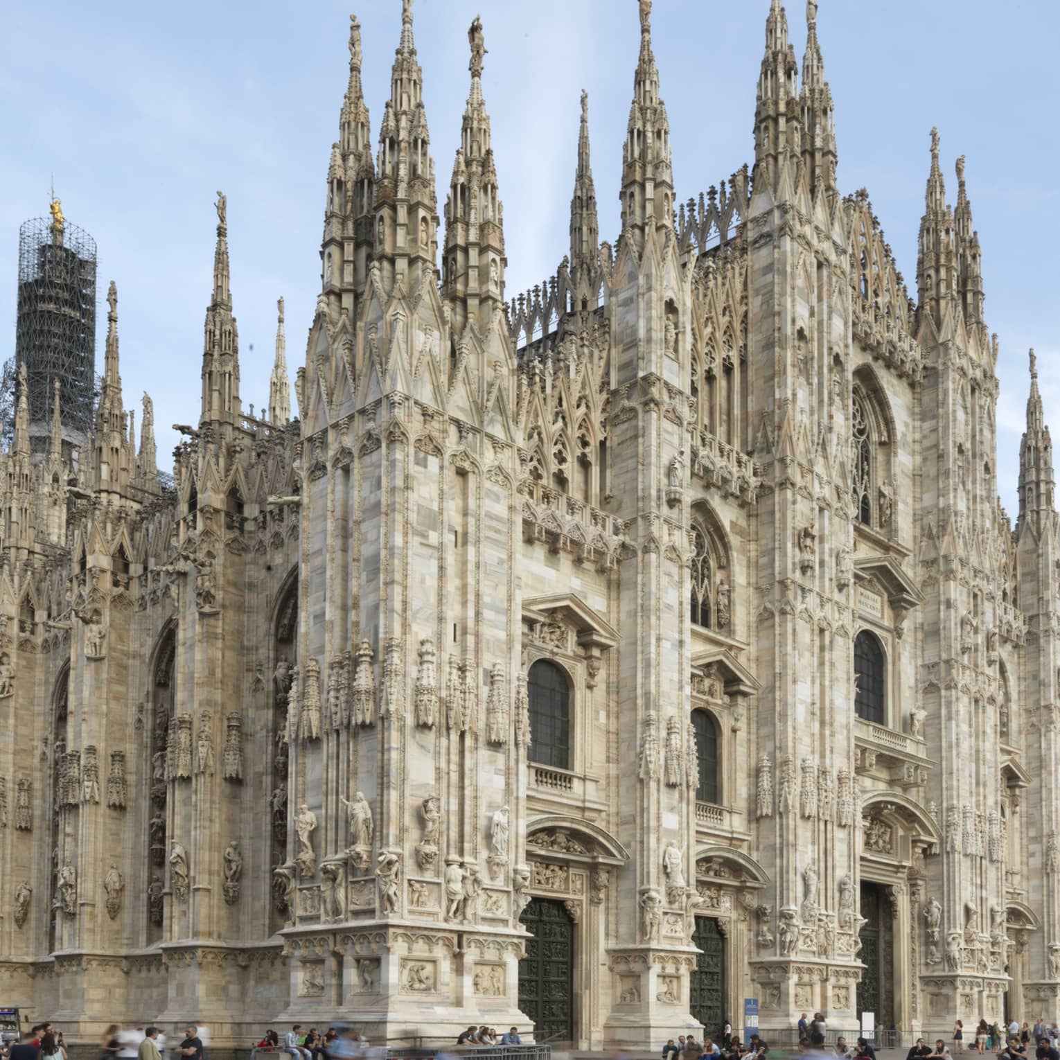 The exterior of the Milan Cathedral (Duomo di Milano), showcasing its intricate Gothic architecture with tall spires and detailed stone carvings against a clear sky.