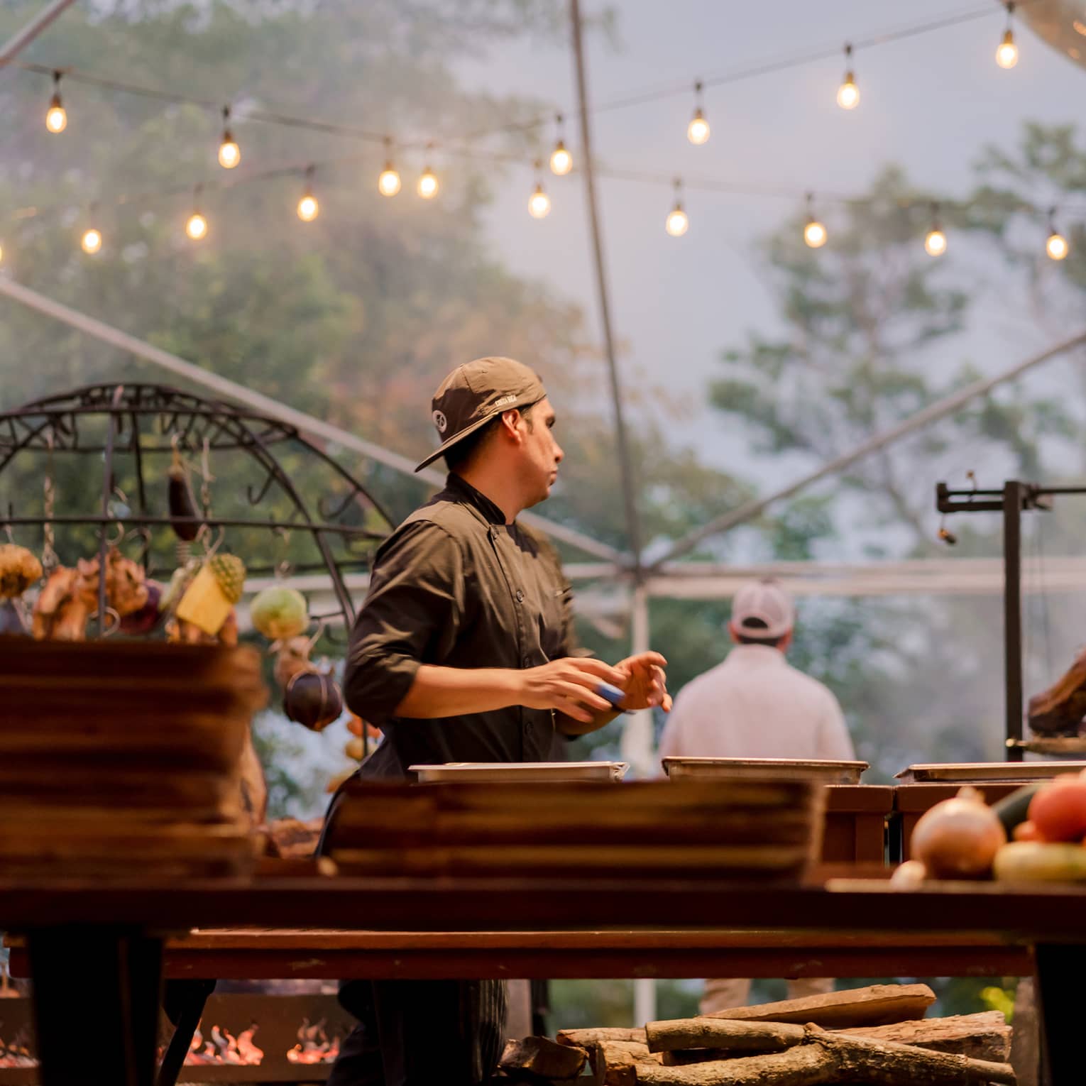 Chef wearing a black chef's jacket stands a grilling station beneath two strings of twinkling lights