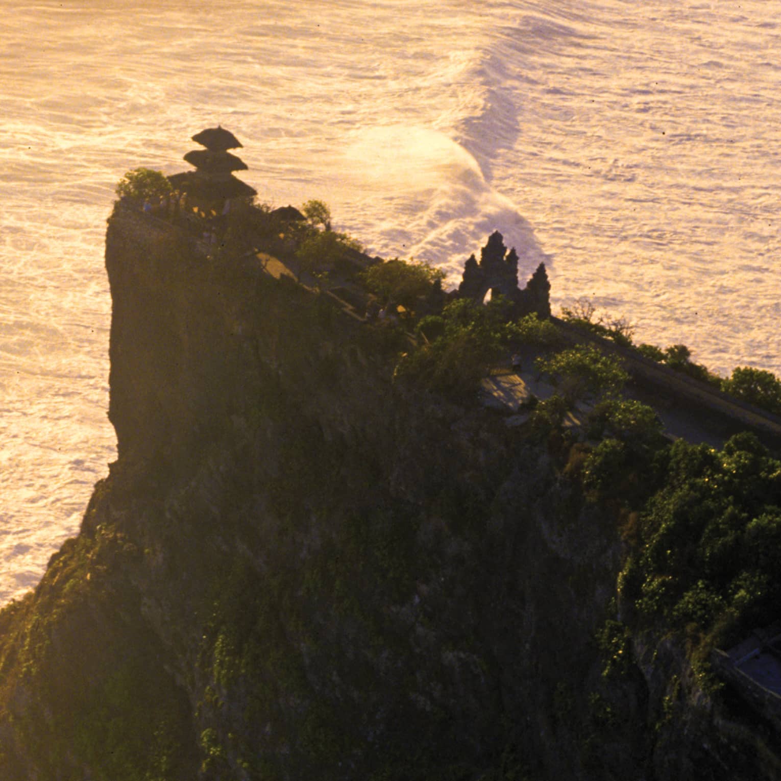 Aerial view of a temple nestled among trees at the edge of a tall, steep cliff; big, white waves crash in the vast sea below.