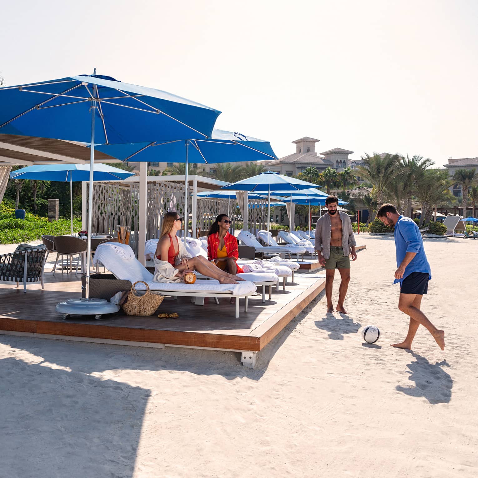 Two beachgoers play soccer while two others sit on lounge chairs, sheltered under blue umbrellas on a beach cabana platform.
