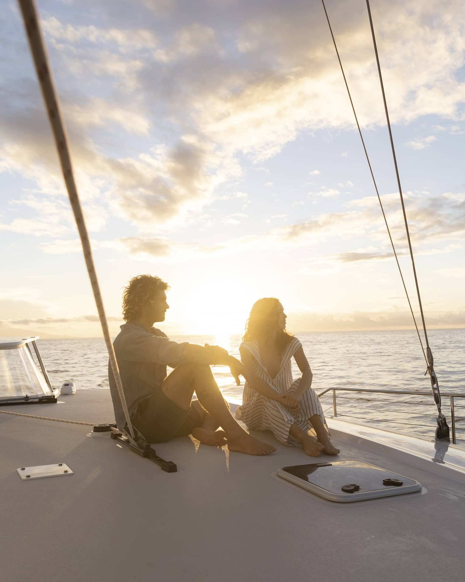 Couple in silhouette sitting barefoot on a boat deck framed by rigging ropes against a bright blue clouded sky at sunset.