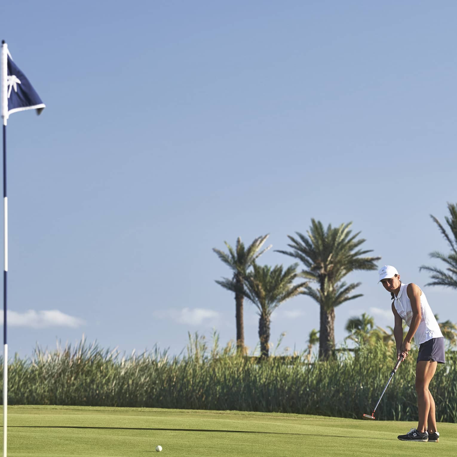 On a bumpy green bordered by reeds and palm trees, a golfer watches the ball's movement toward a hole marked by a blue flag. 