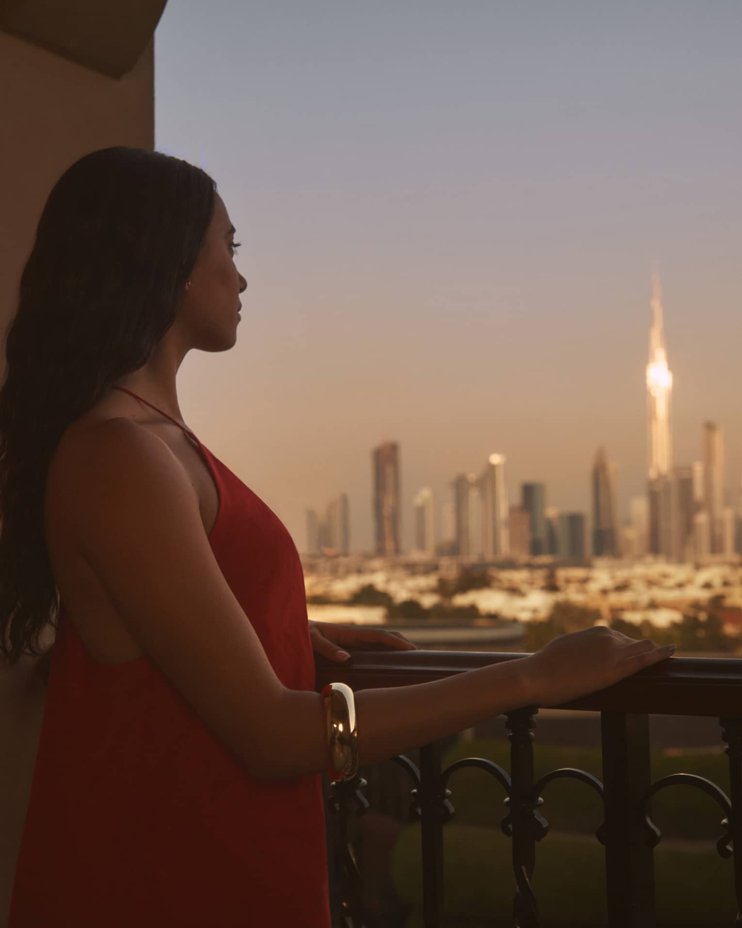 Person with long brown hair standing on balcony with hand resting on railing, looking out to a city at dusk