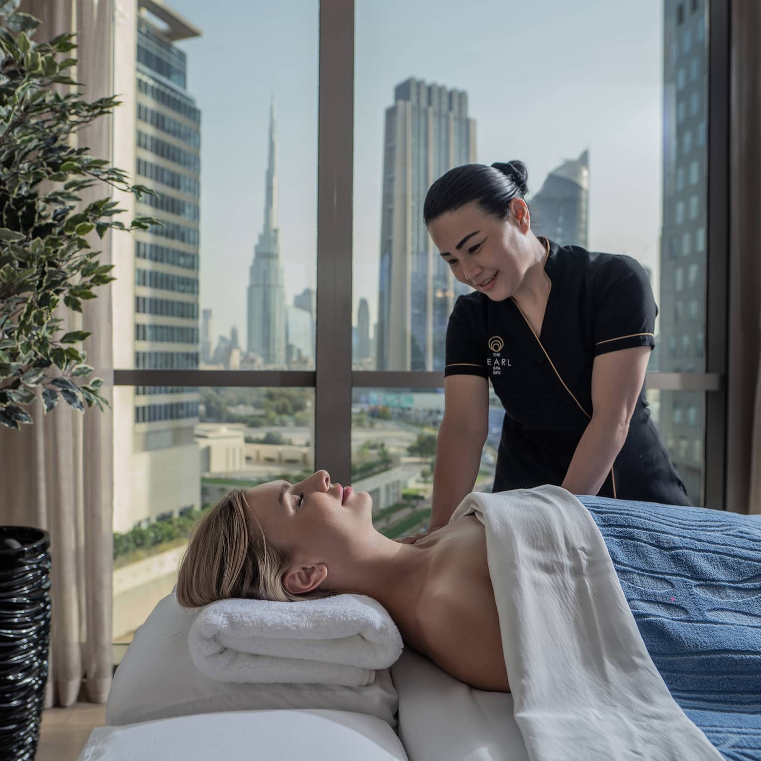A guest enjoys a massage from a Hotel employee. Behind them, a window frames tall glass skyscrapers and clusters of trees.